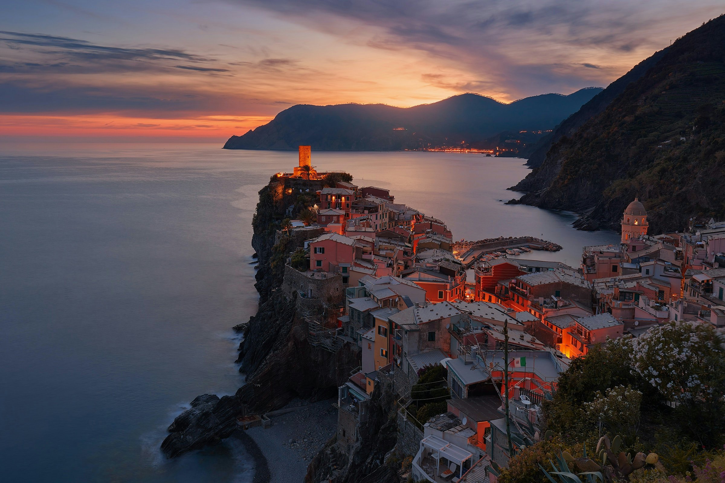 Vernazza's clifftop houses and harbour tower glow orange at sunset in Italy's Cinque Terre.