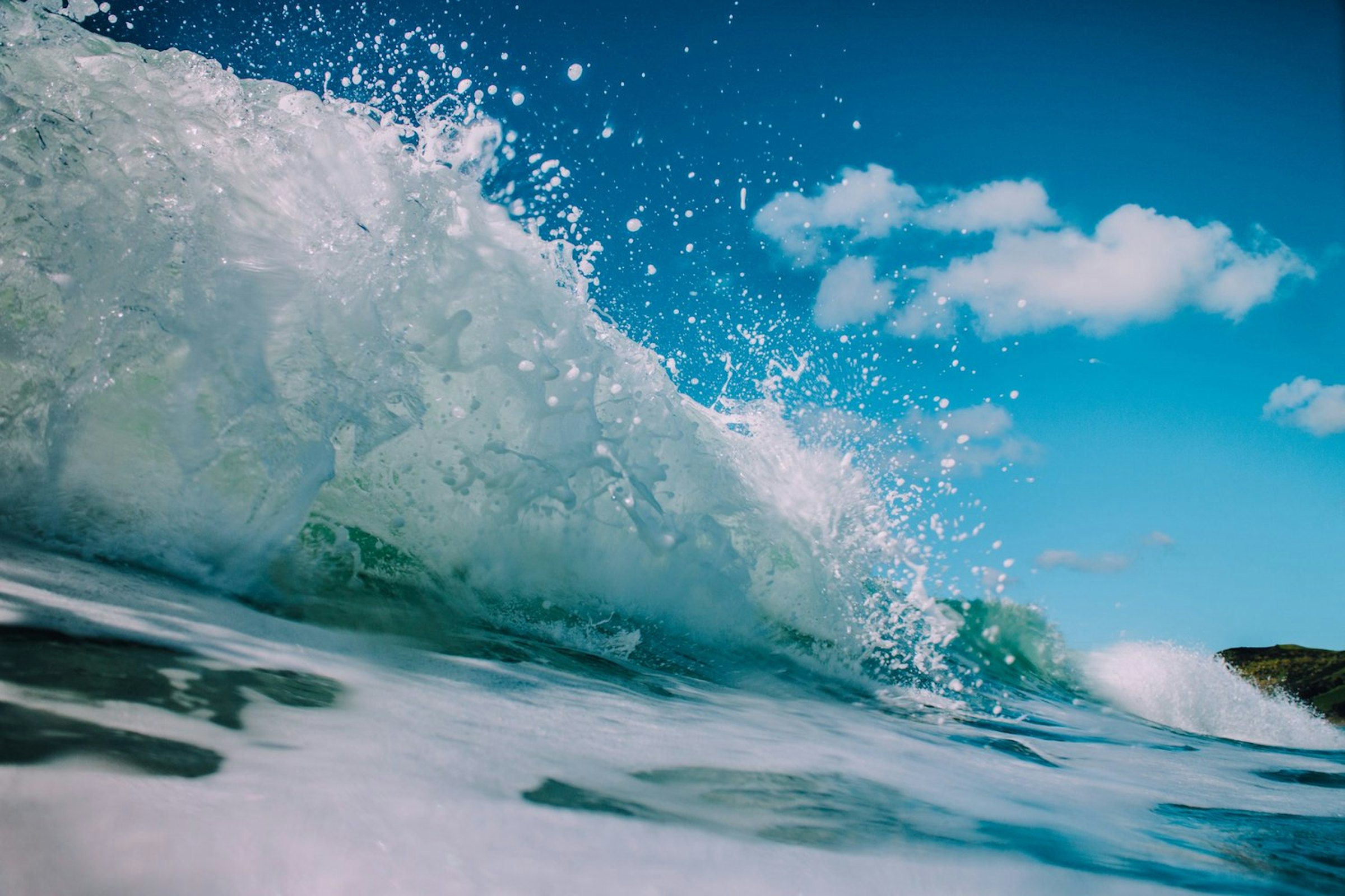 A turquoise wave curls and breaks into white spray under a clear blue sky.