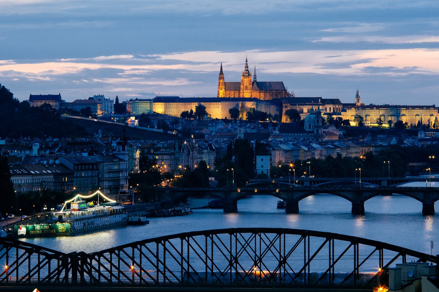 czech republic by train a view of the Prague castle on the hill lit up in the evening sky, with the bridges over the river and cityscape in the foreground