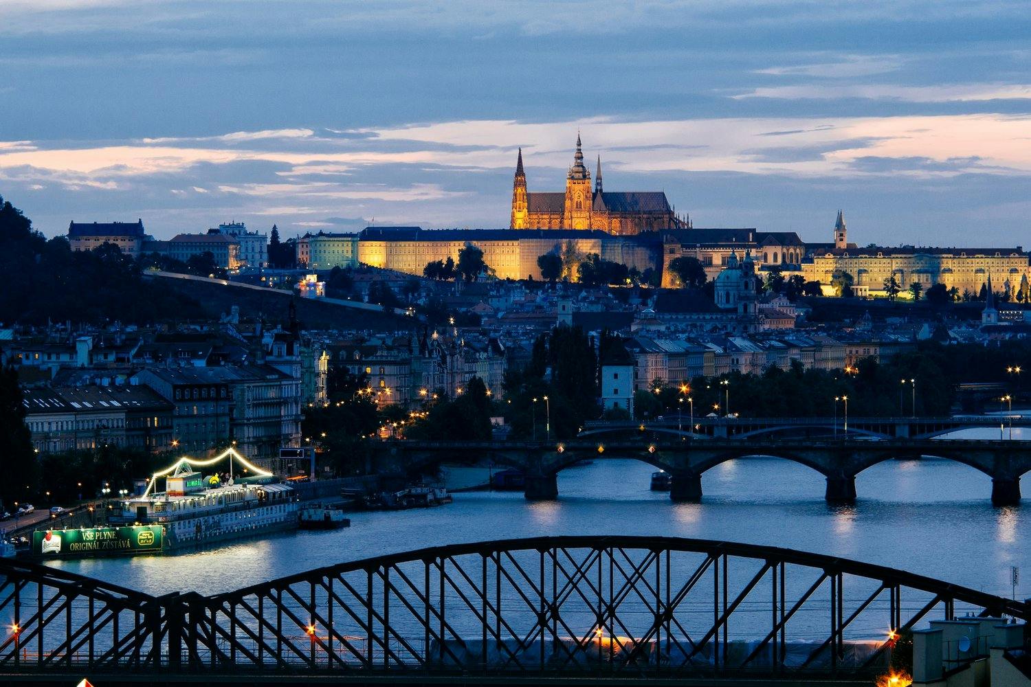 a view of the Prague castle on the hill lit up in the evening sky, with the bridges over the river and cityscape in the foreground