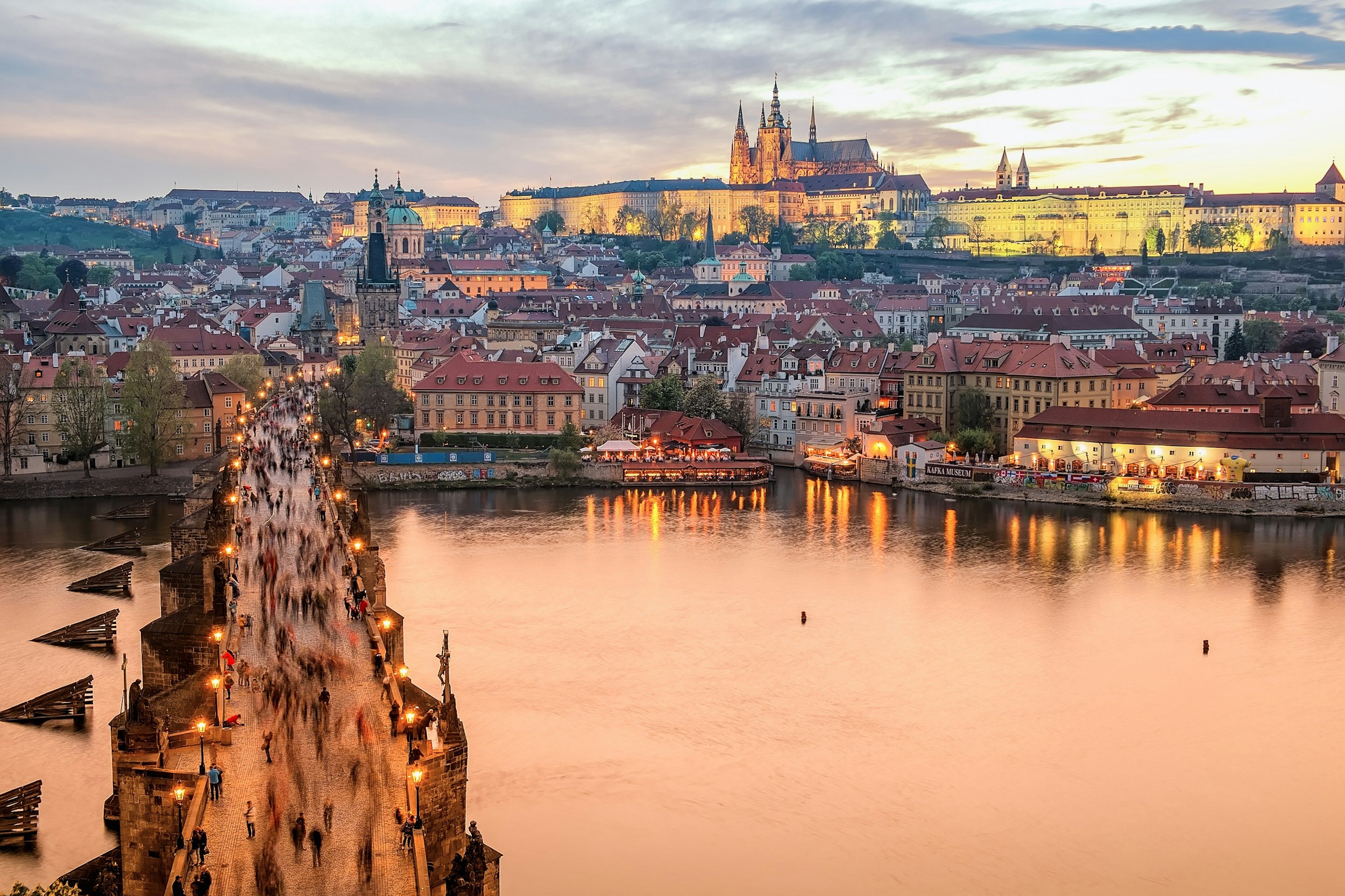 aerial view of the charles bridge in prague with people walking over it, with the medieval buildings of prague in the background as well as the castle on top of the hill