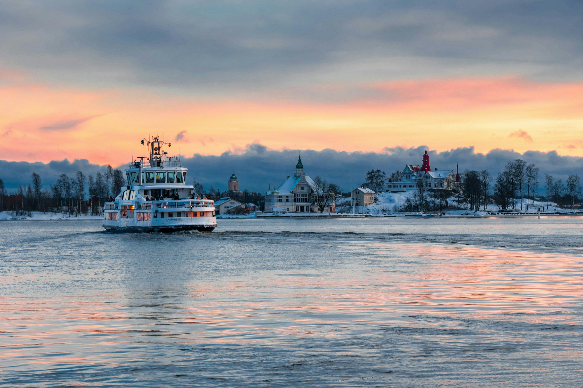 Ferry crossing calm water past a snow-dusted coastal town with domed buildings, dusk sky, Finland