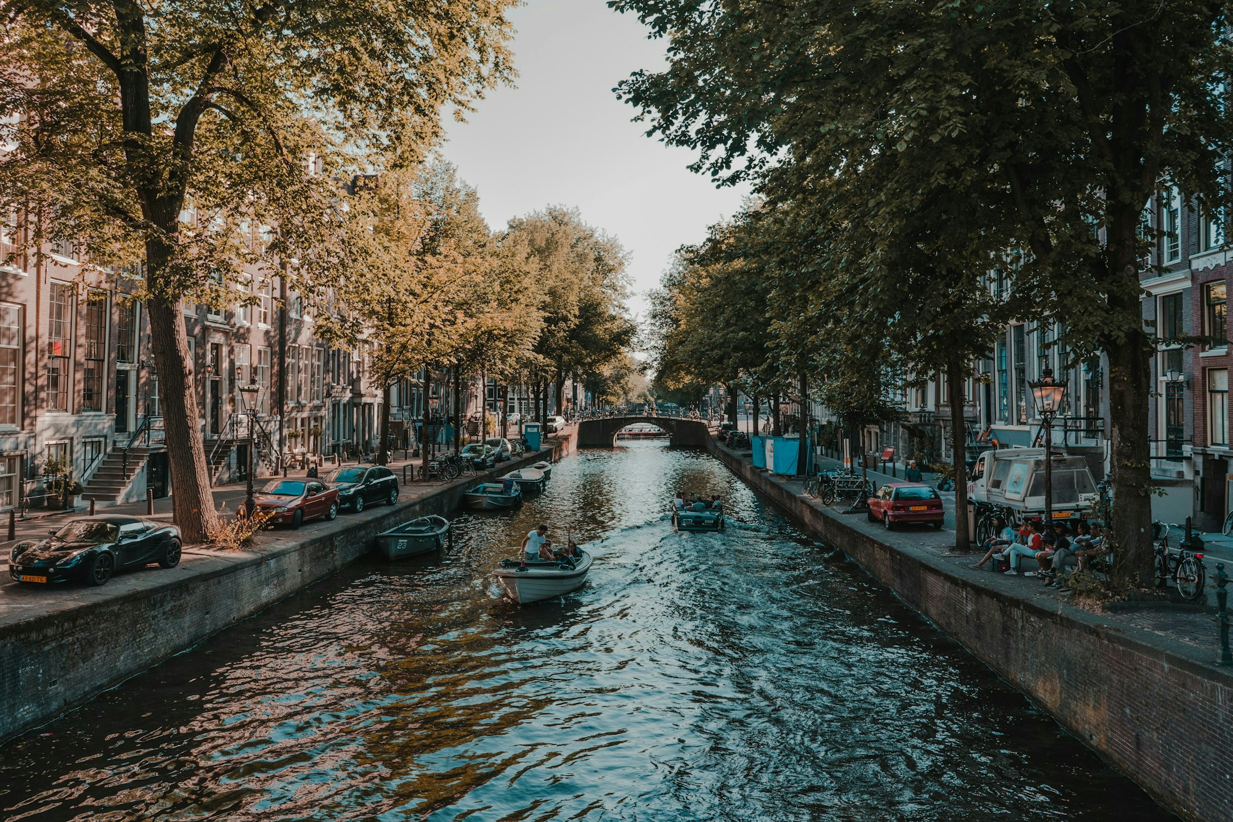 Small boats cruise a tree-lined Amsterdam canal flanked by townhouses in warm afternoon light.