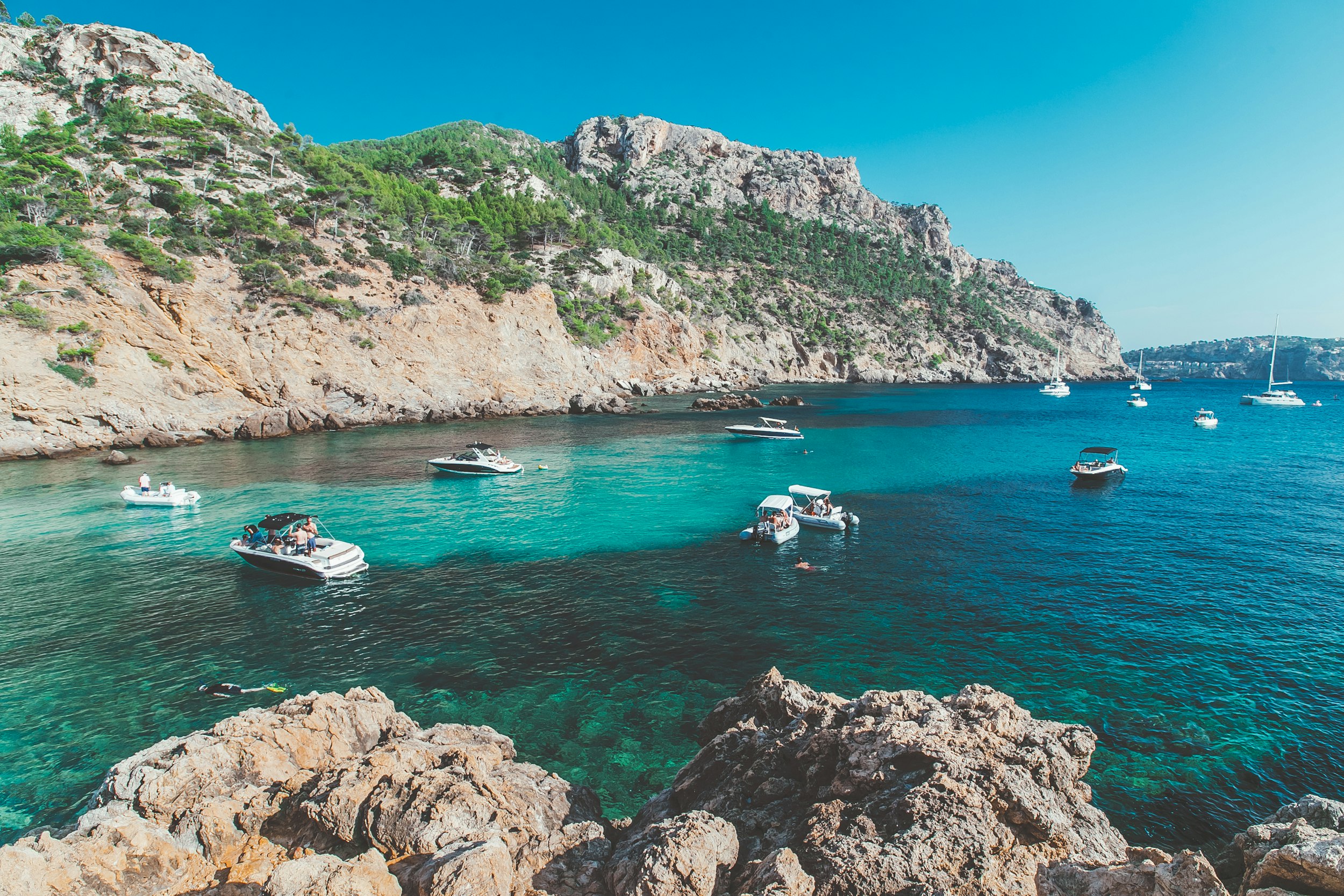 white boats in an azure sea bay in Mallorca, with rocky and green cliffs in the background, during daytime