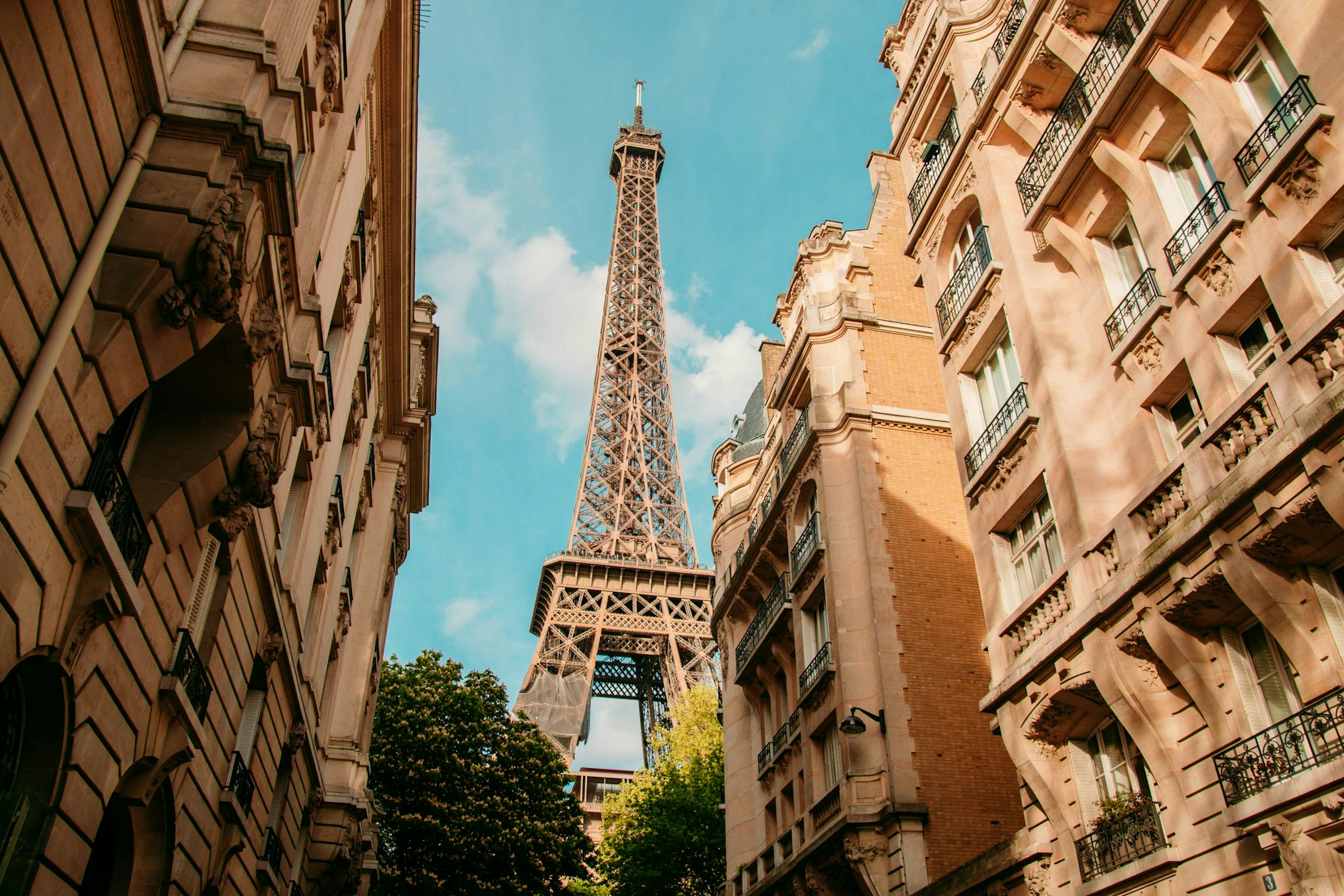 The Eiffel Tower rises above Parisian apartment buildings, seen from a narrow street below.