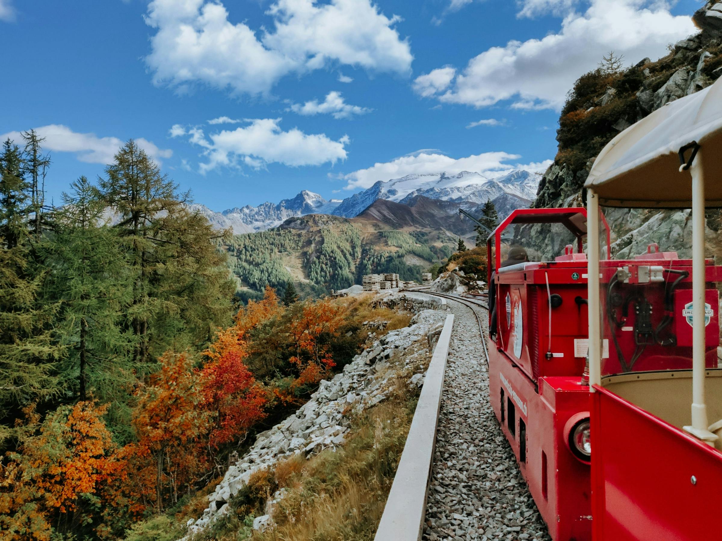 A red narrow-gauge train winds past autumn larches, snow-capped Alpine peaks rising ahead.