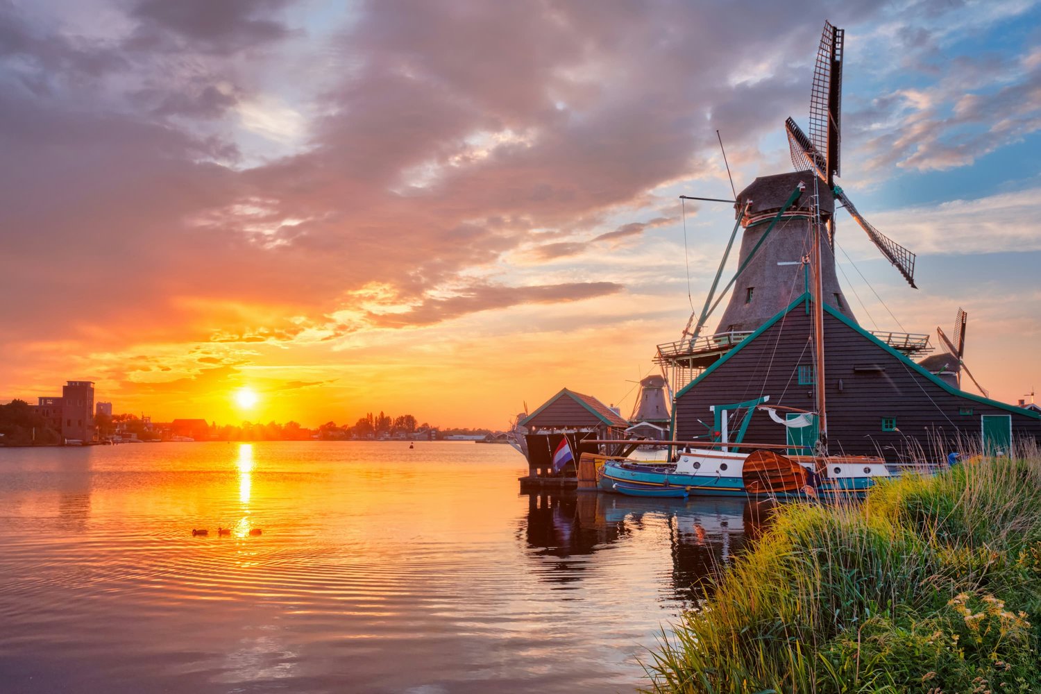 A sunset over the Dutch river at Kinderdijk in the Netherlands, showing one of the famous windmills and a little cabin and fishing boat in front of it, in the background showing the city skyline.