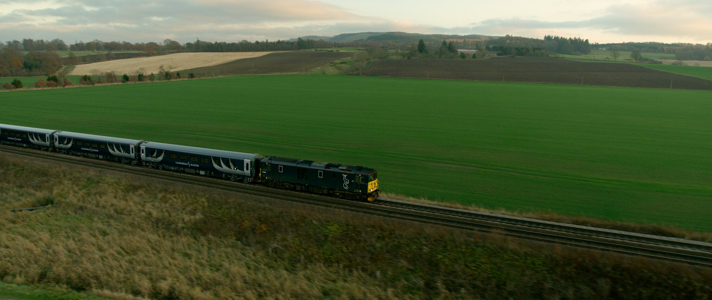 The Caledonian Sleeper train crossing green Scottish farmland, seen from above at dusk.