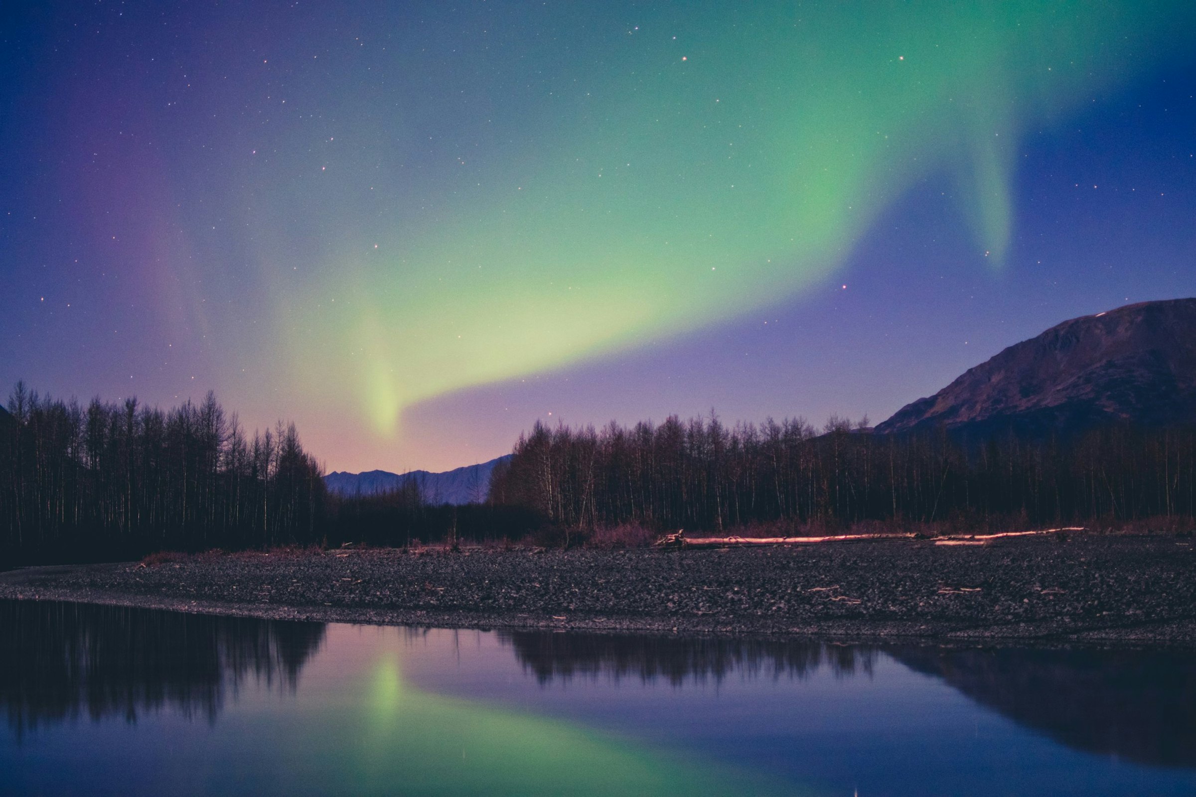 Green and purple northern lights ripple above a forest lake reflecting the night sky.