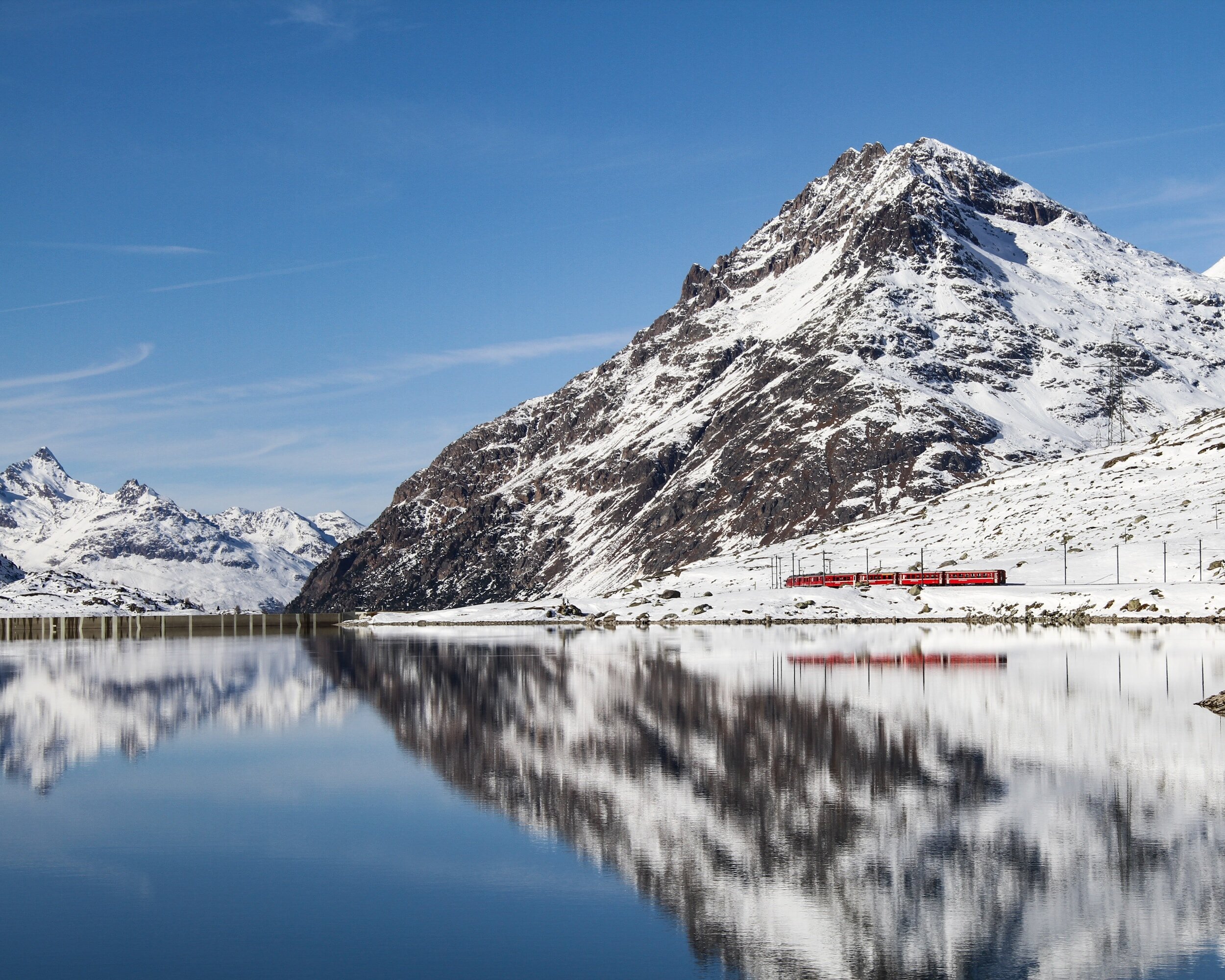 a red train in front of a white snowy mountain with blue skies, and the same picture reflected in the lake in front of it