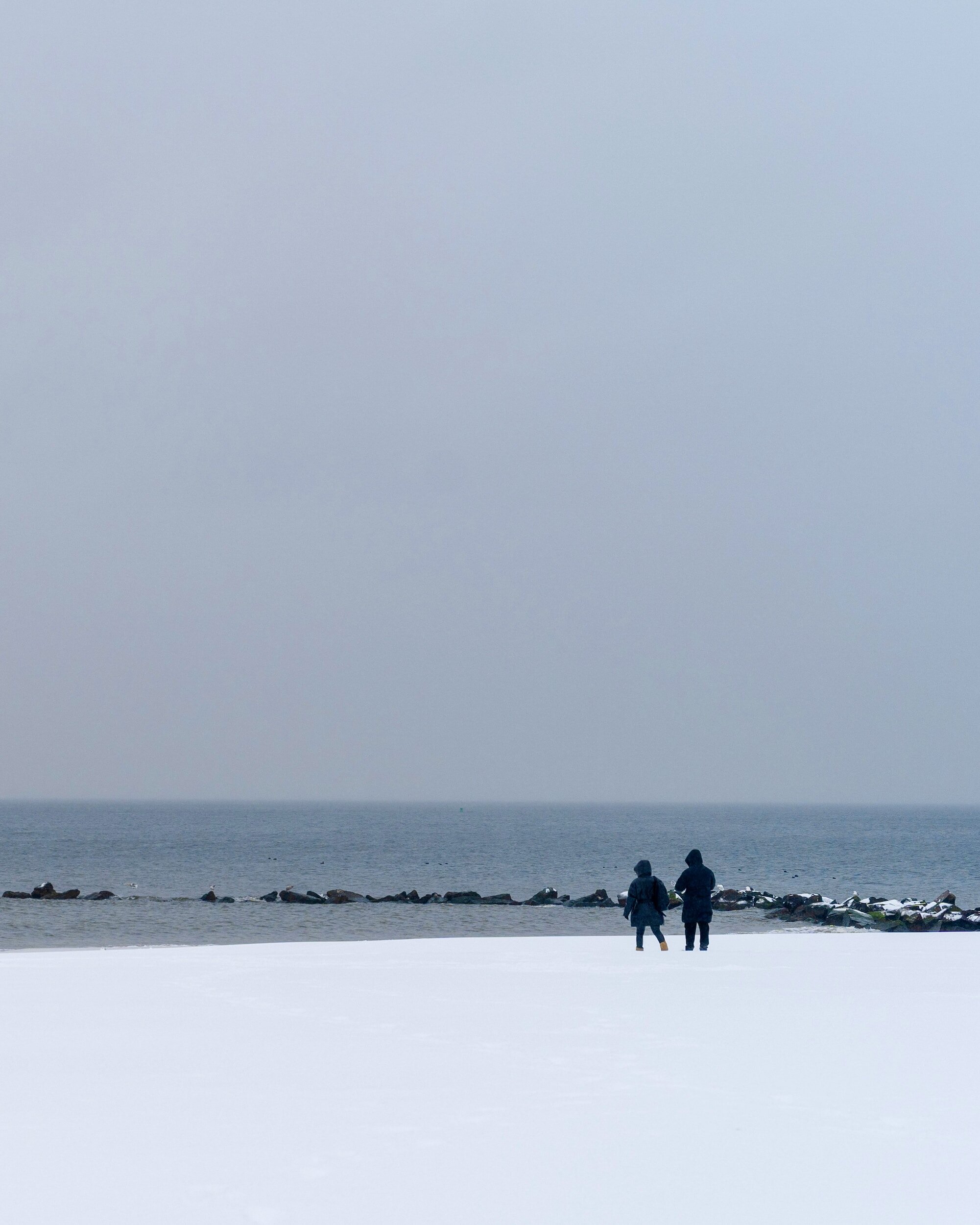 a white snowy beach with a blue sea and grey sky, two people walking in winter clothes