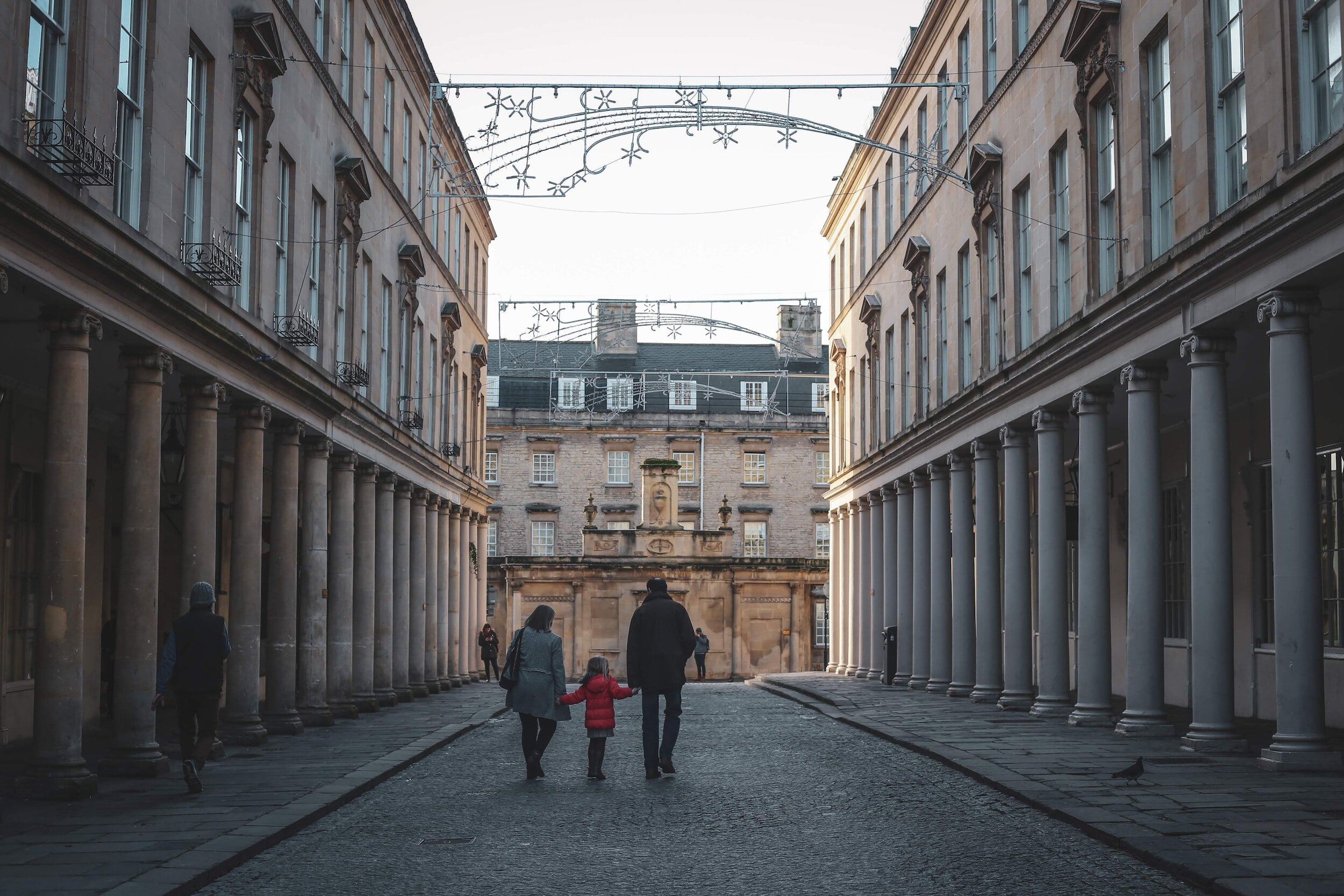 a couple and their child walking in the city of Bath, between the buildings in roman architecture, with christmas lights at the top.