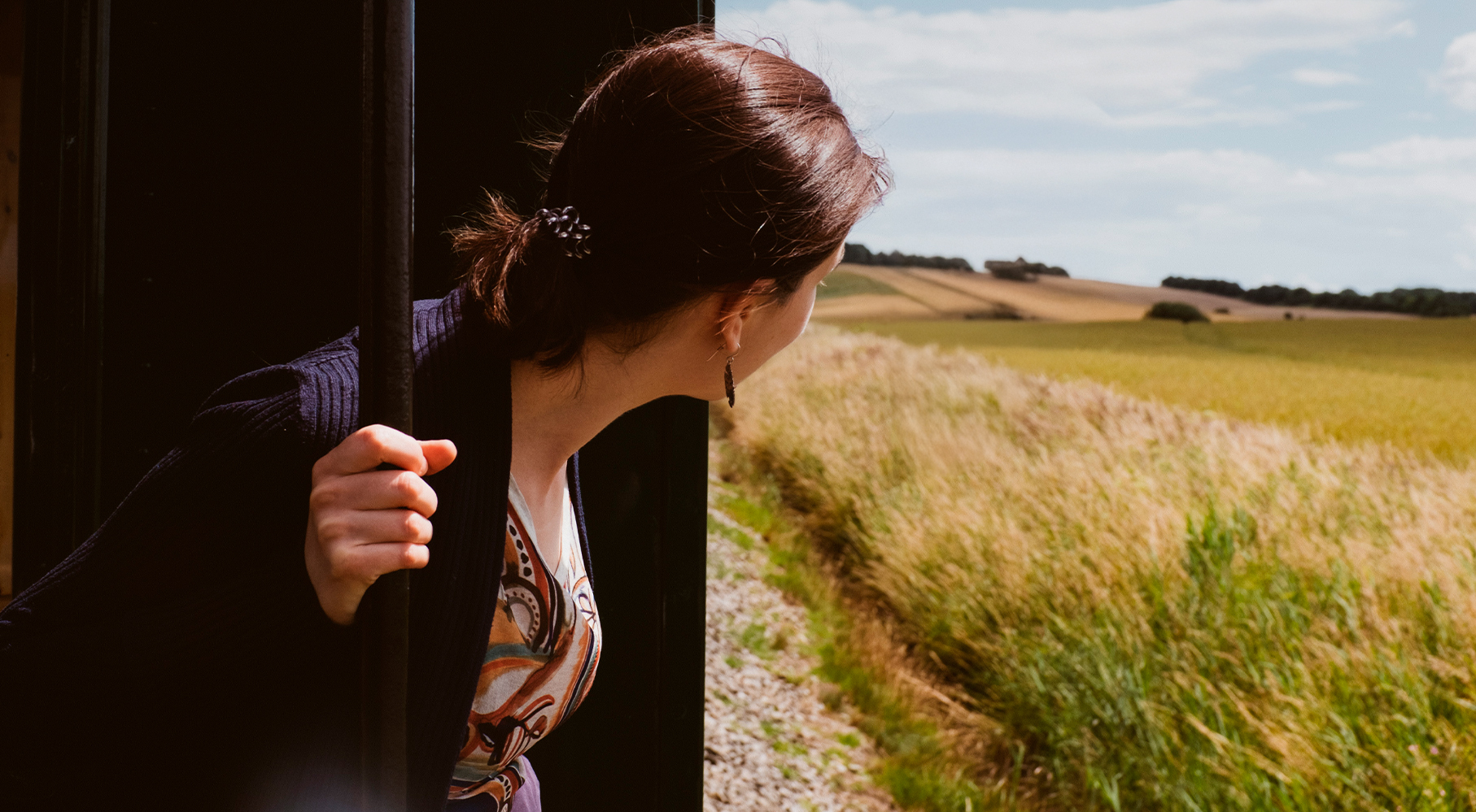 Woman leaning out of a train facing away from the camera, looking out at the scenic rural landscape.