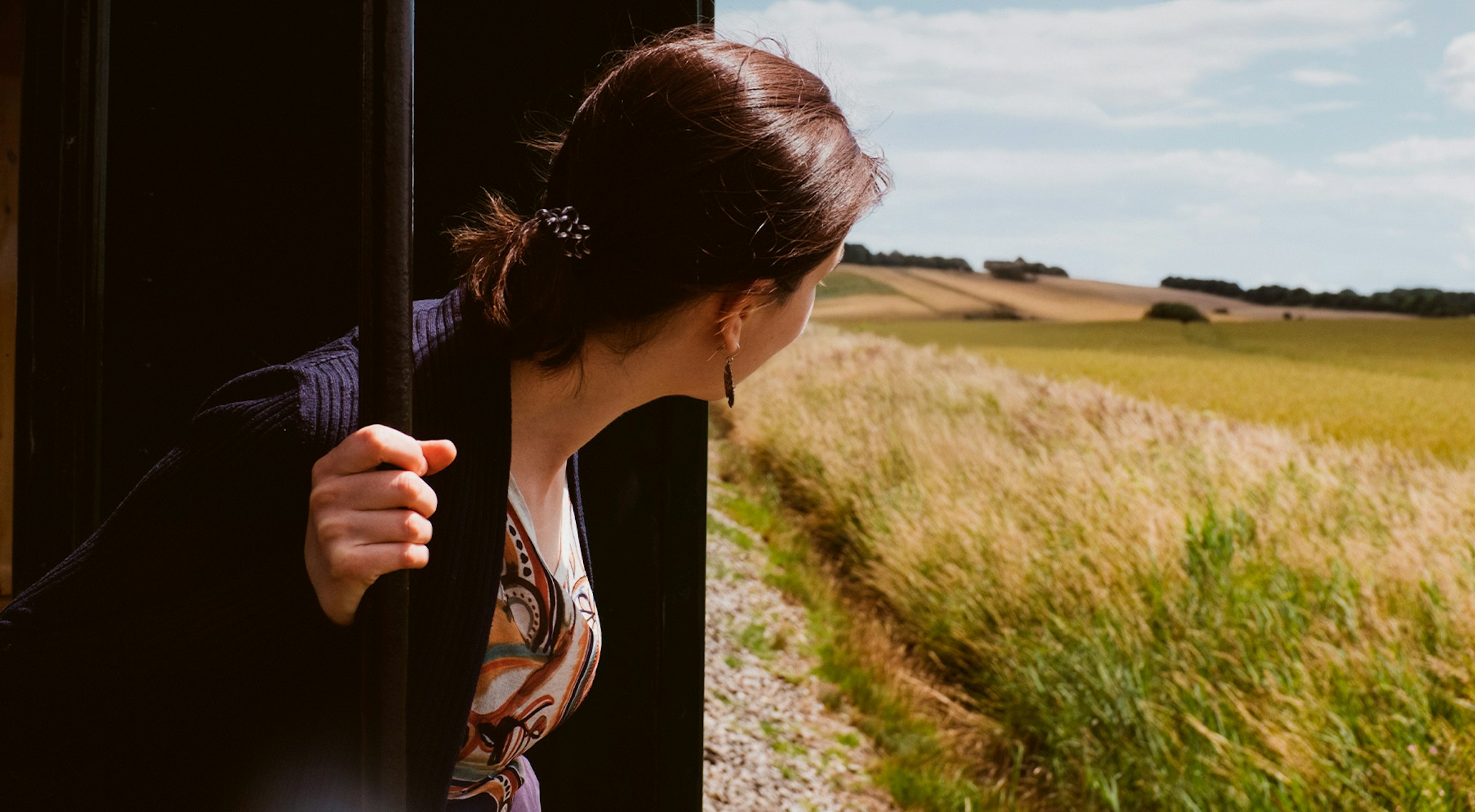 Woman leans out of an open train doorway over golden fields under a partly cloudy sky.