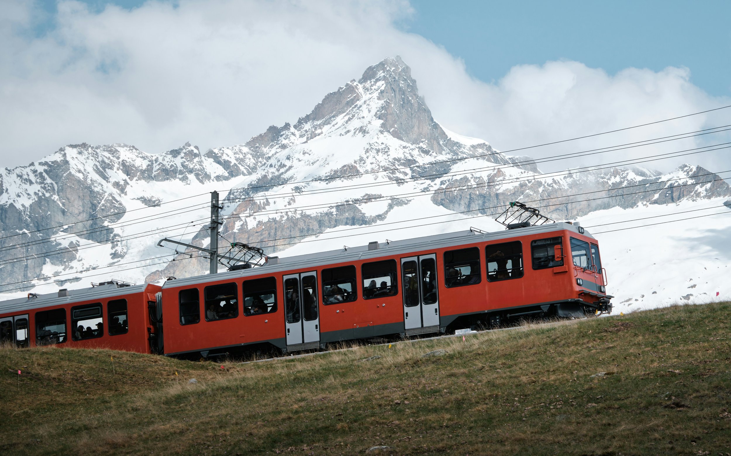 A red train going up a hill in Switzerland, with snowy peaks behind it
