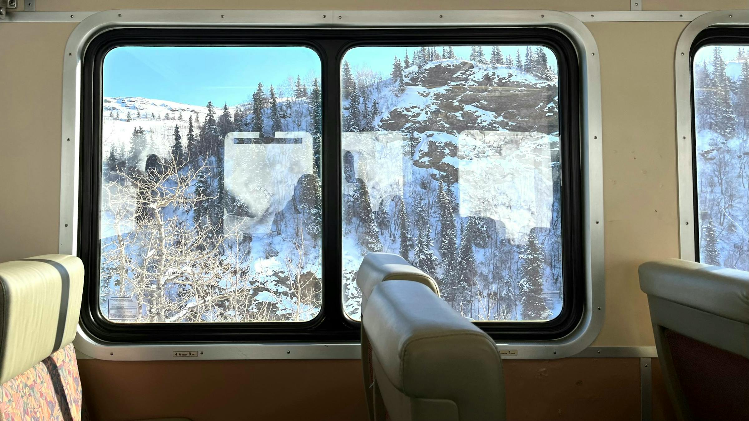 Snow-covered mountains and pine forest seen through a train window, cream seats in foreground.