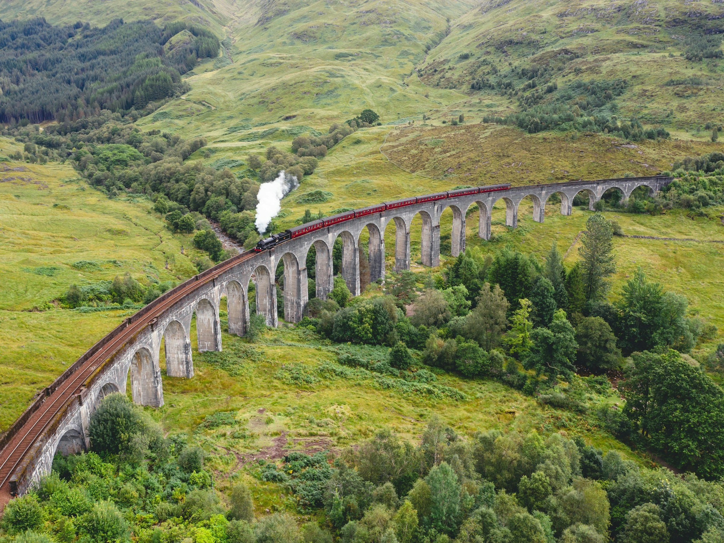 A steam train crosses the curved Glenfinnan Viaduct in the Scottish Highlands, aerial view.