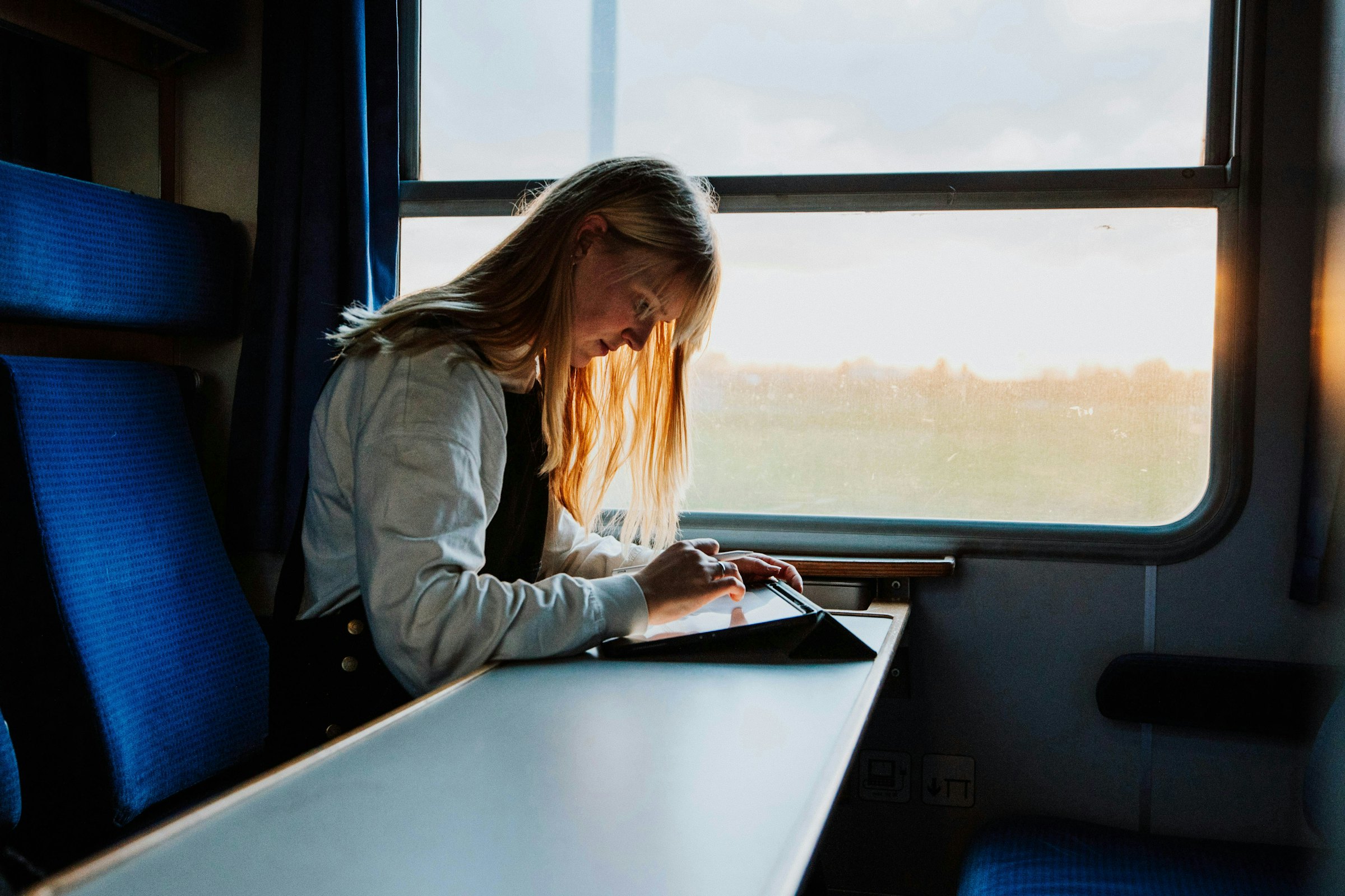 Woman using a tablet at a train window table, lit by warm evening sunlight.