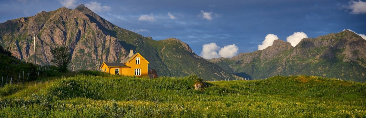 A yellow house sits atop a green valley with grey moutains in the background, in Norway.
