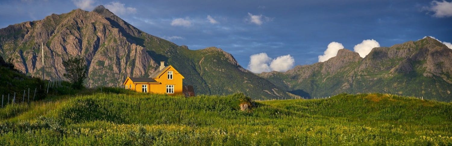 A yellow house sits atop a green valley with grey moutains in the background, in Norway.