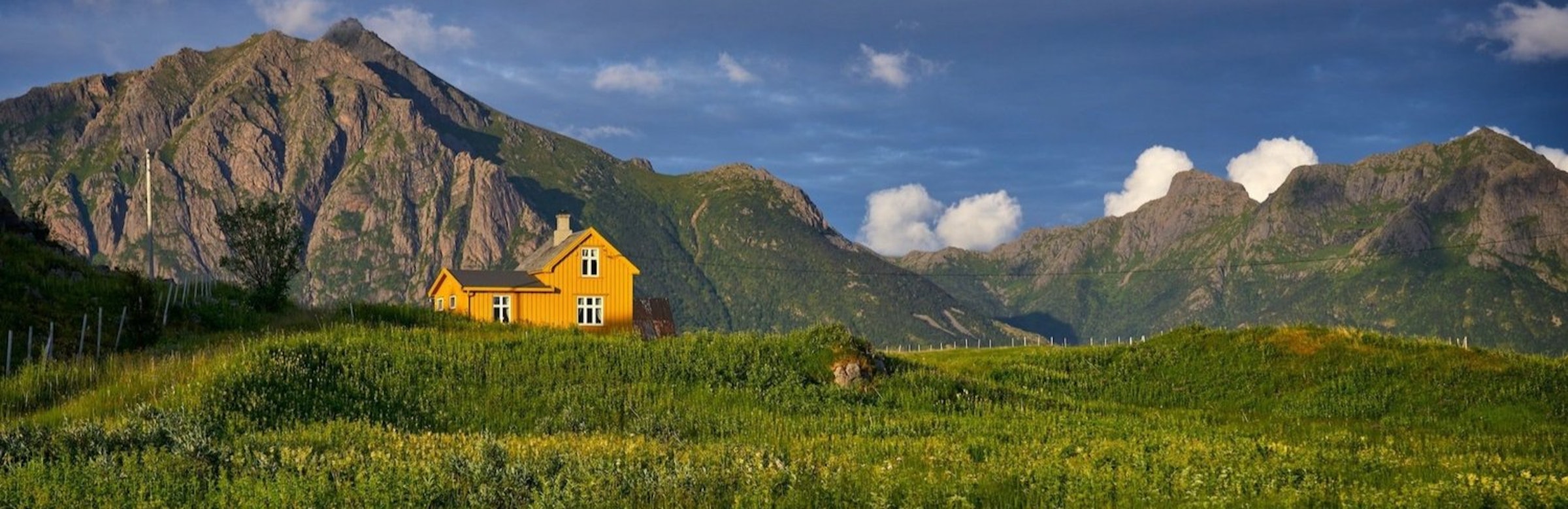 Een geel huis staat bovenaan een groene vallei met grijze bergen op de achtergrond, in Noorwegen.