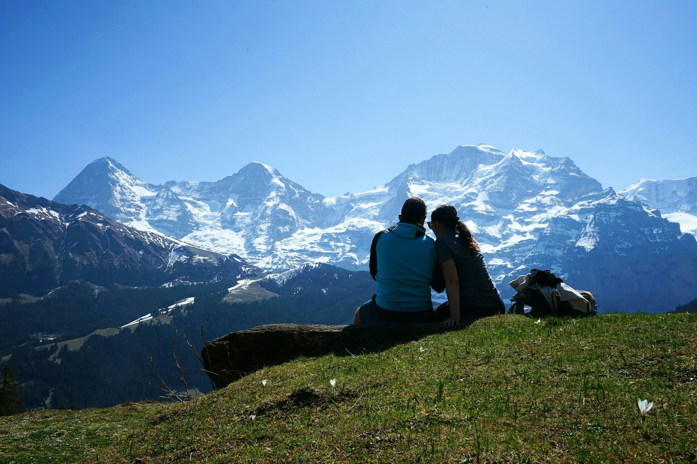 A couple sits together on a rock in a flower-dotted alpine meadow, facing snow-capped mountain peaks.