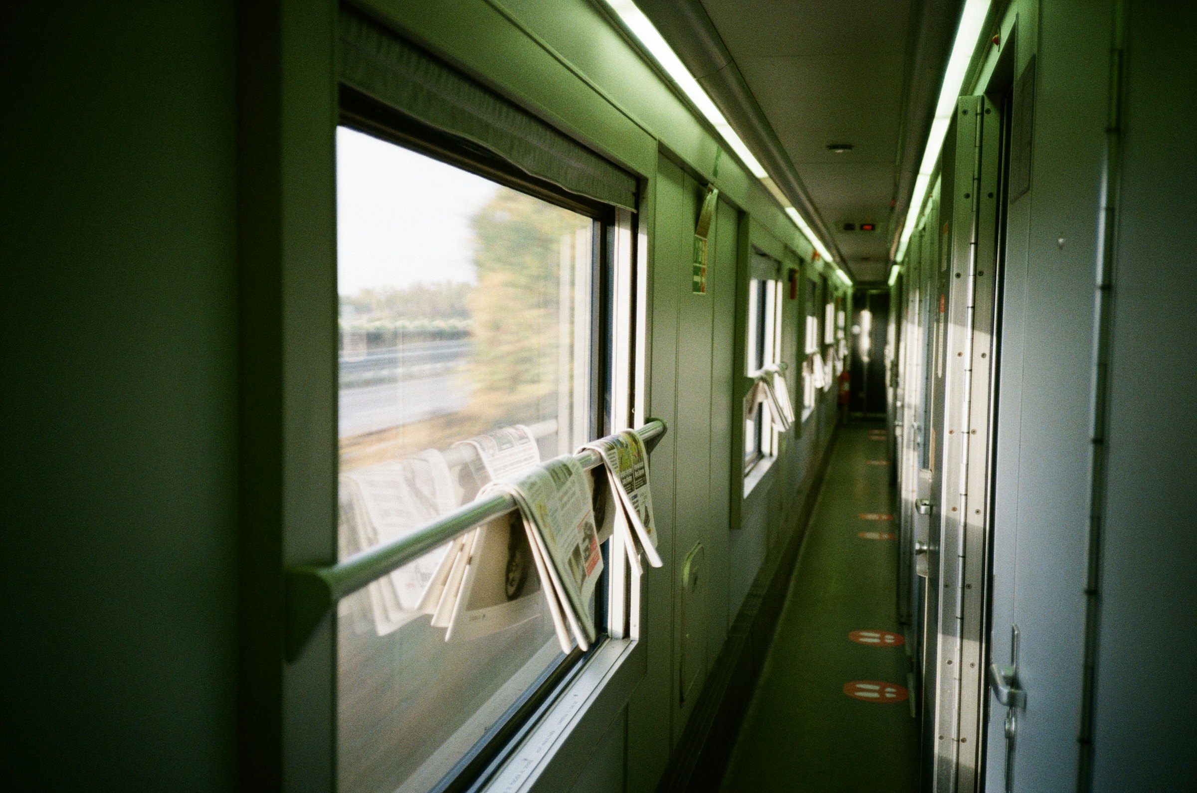 Sunlit corridor of a European sleeper train carriage, with newspapers tucked along the handrail.