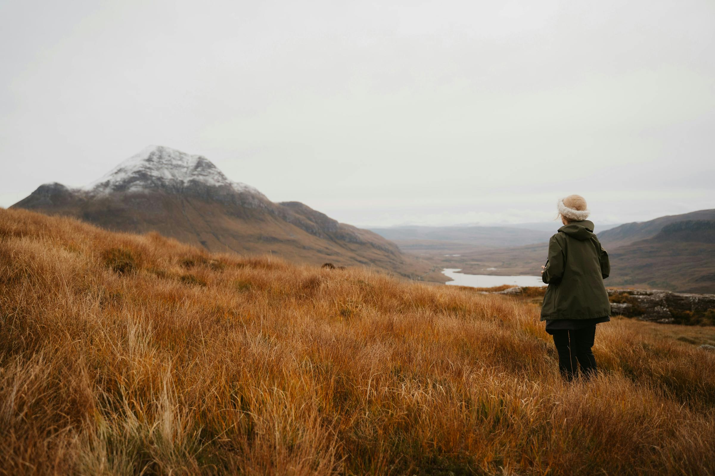 A solo traveller in a wool hat looks across autumn moorland toward a snow-dusted highland peak.