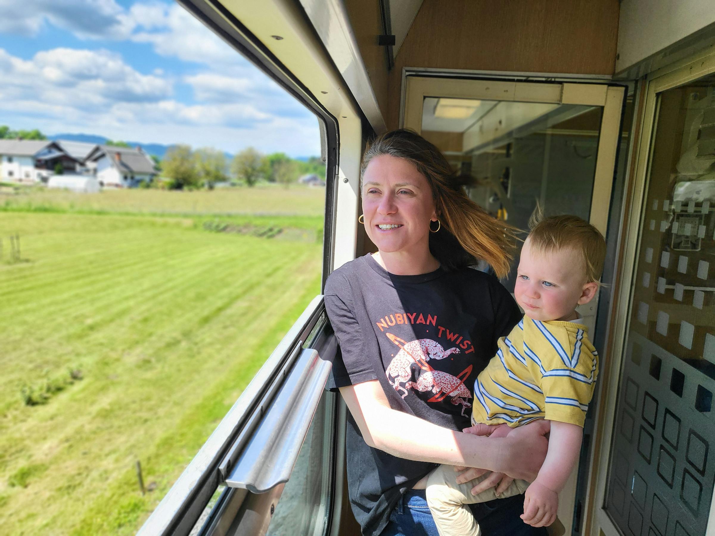 Jo & Ellis taking in the view as they snake through the Austrian countryside. A mother holds her toddler up to an open train window to watch green countryside roll past.