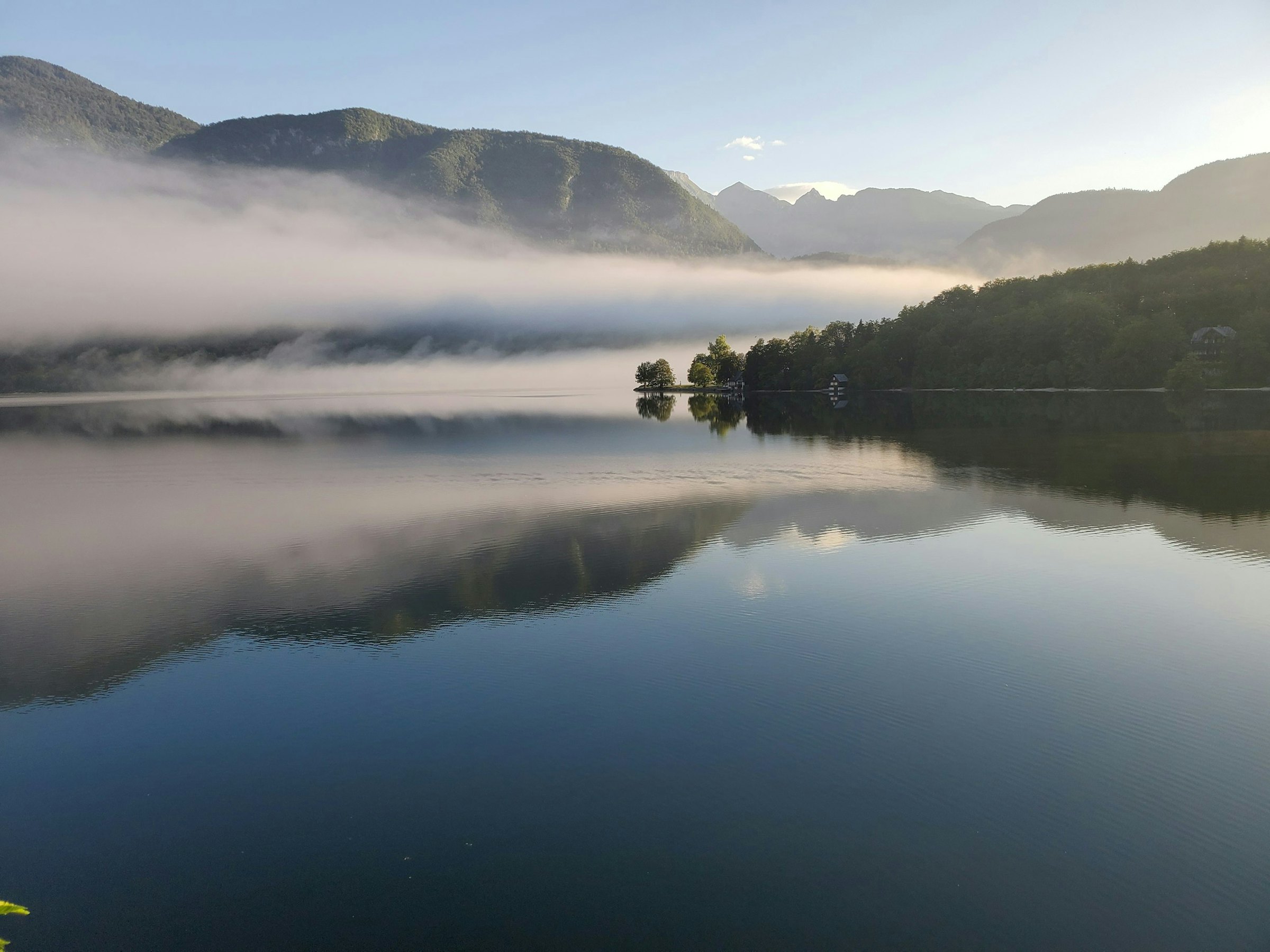 Morning mist drifts over a still alpine lake, reflecting forested slopes and distant mountains.
