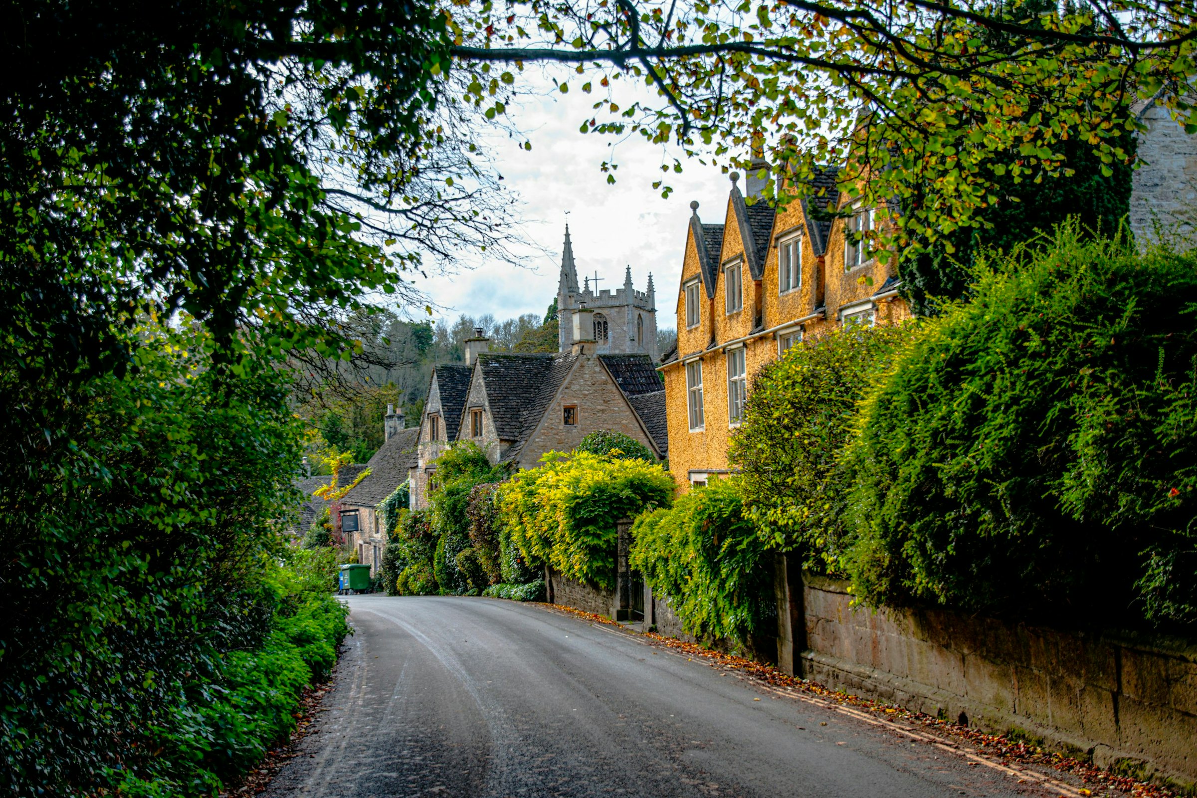 A quiet lane through a Cotswolds-style English village, past honey-stone cottages and a church spire.