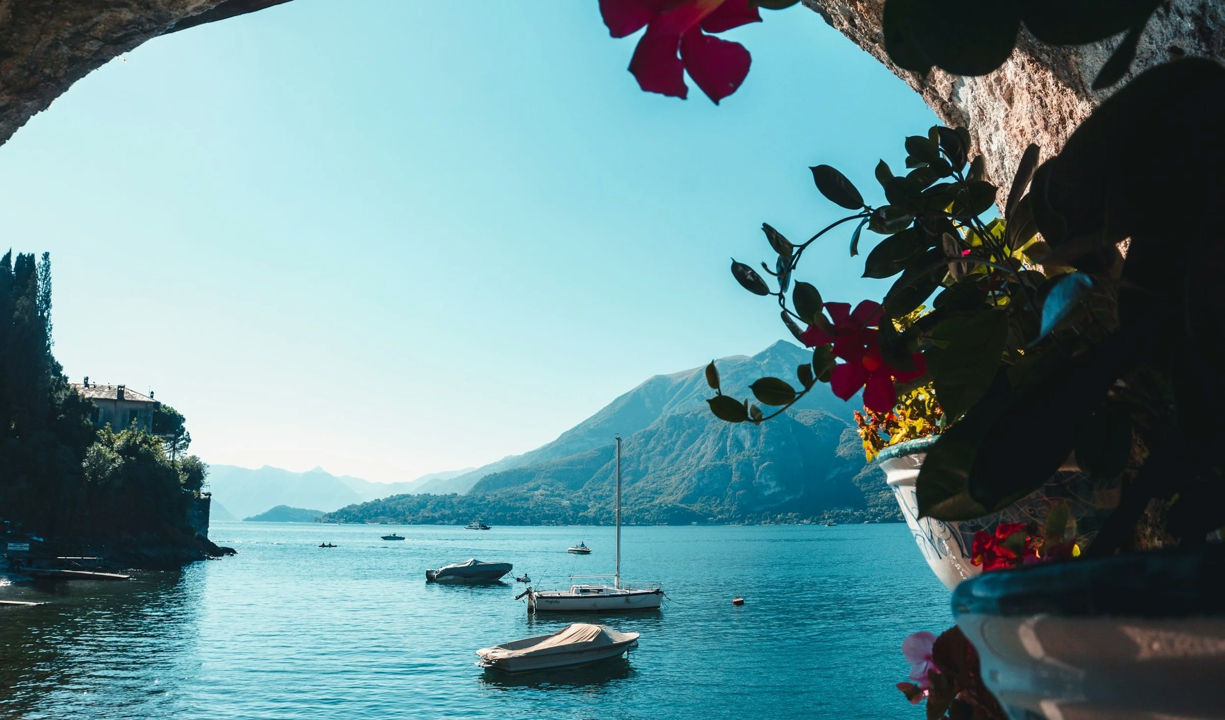 Lake Como framed by an archway of pink flowers, with small boats moored and mountains beyond.
