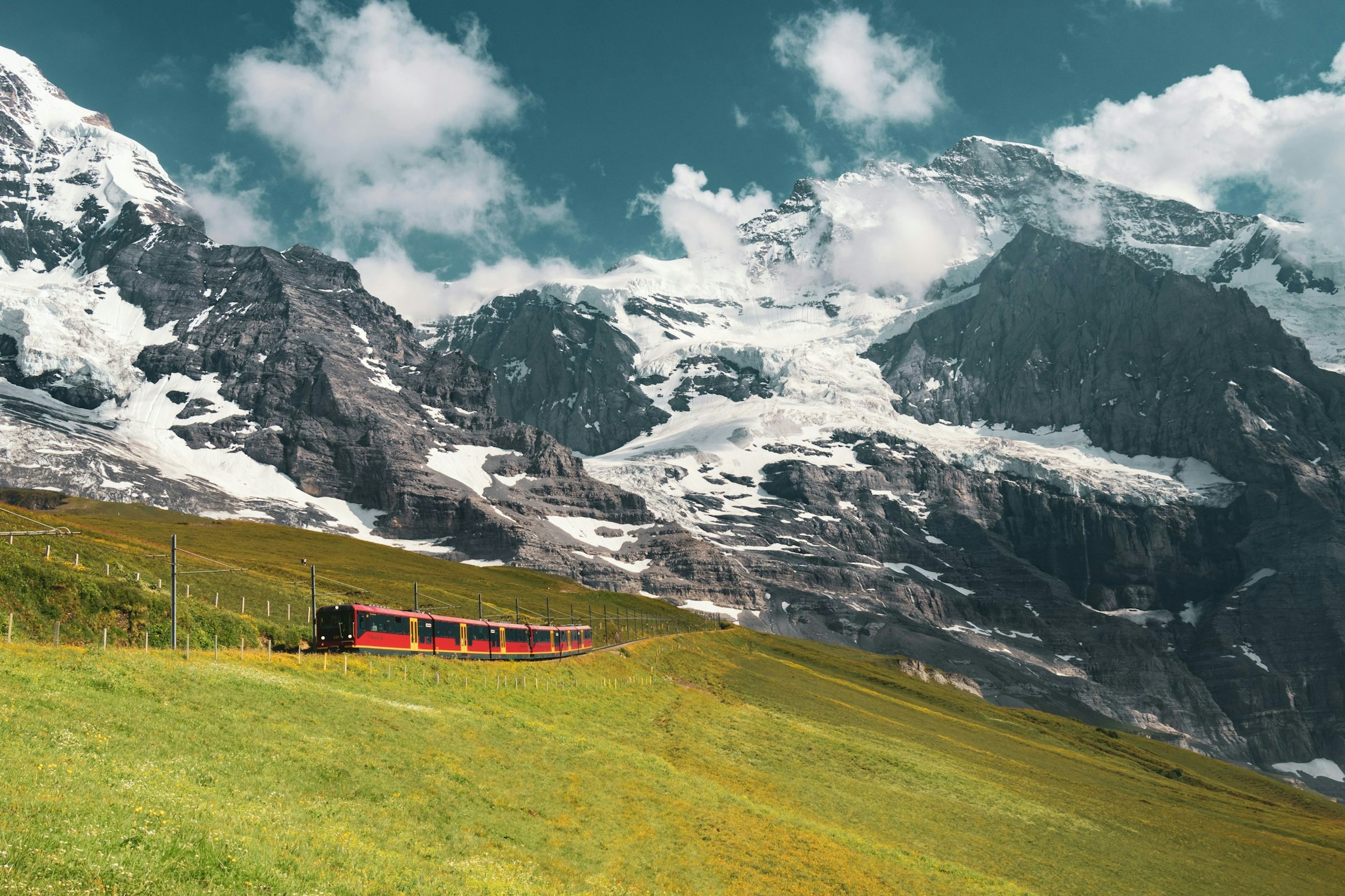 A red mountain train crosses a green alpine slope beneath snow-covered rocky peaks and cloudy sky.