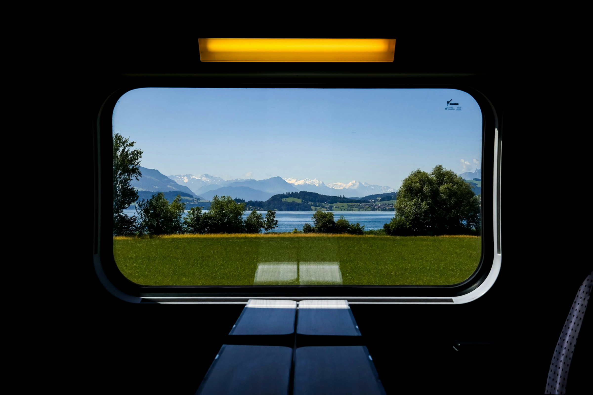 View through a train window of a lake framed by snow-capped mountains and green meadow.