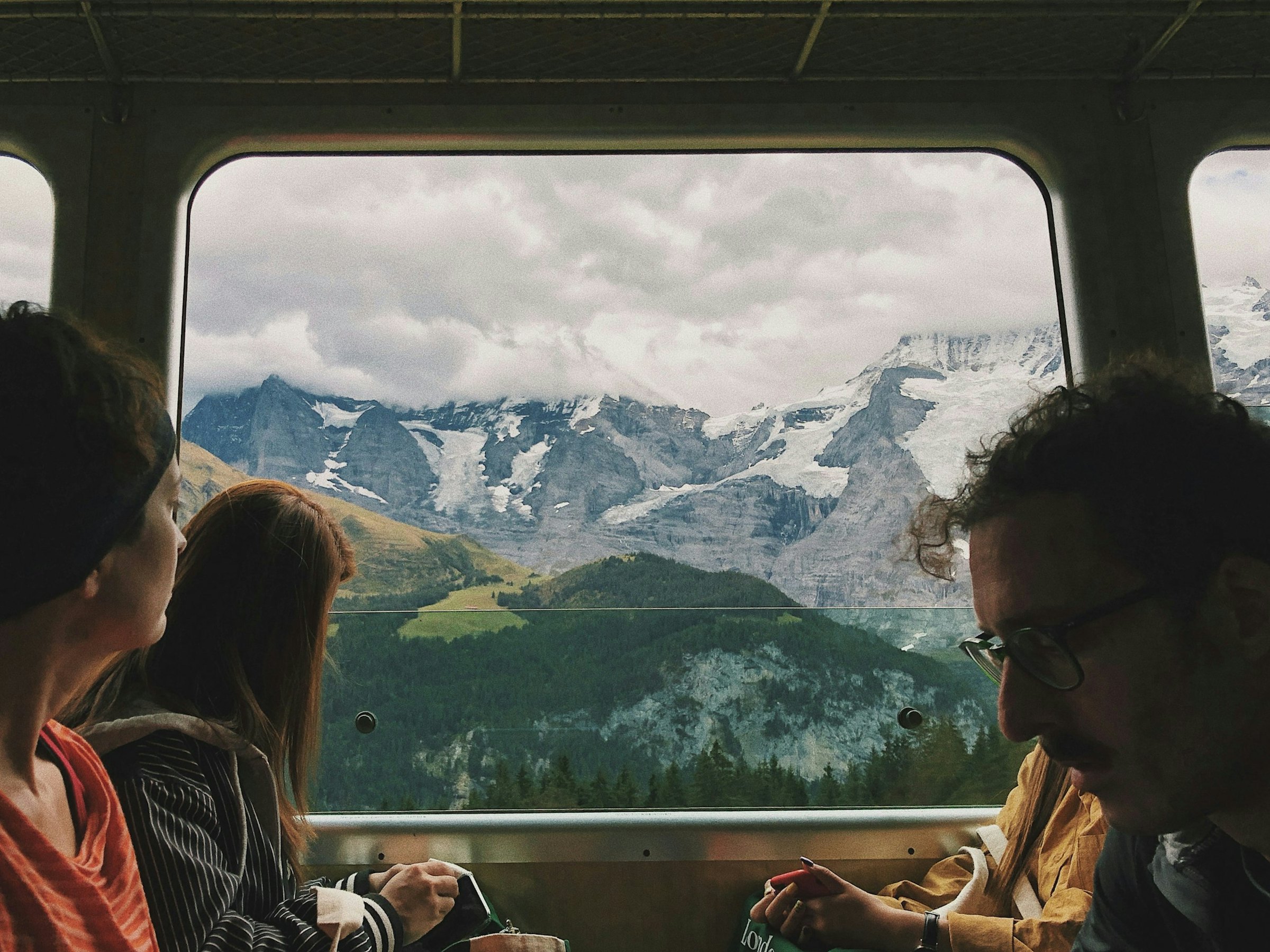 Passengers look out a train window at snow-capped alpine peaks and a forested valley below.