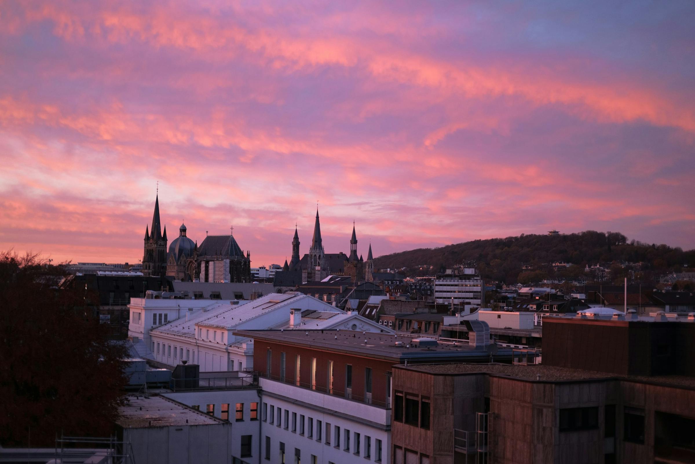 Aachen's cathedral spires stand silhouetted against a vivid pink sunset over the rooftops.