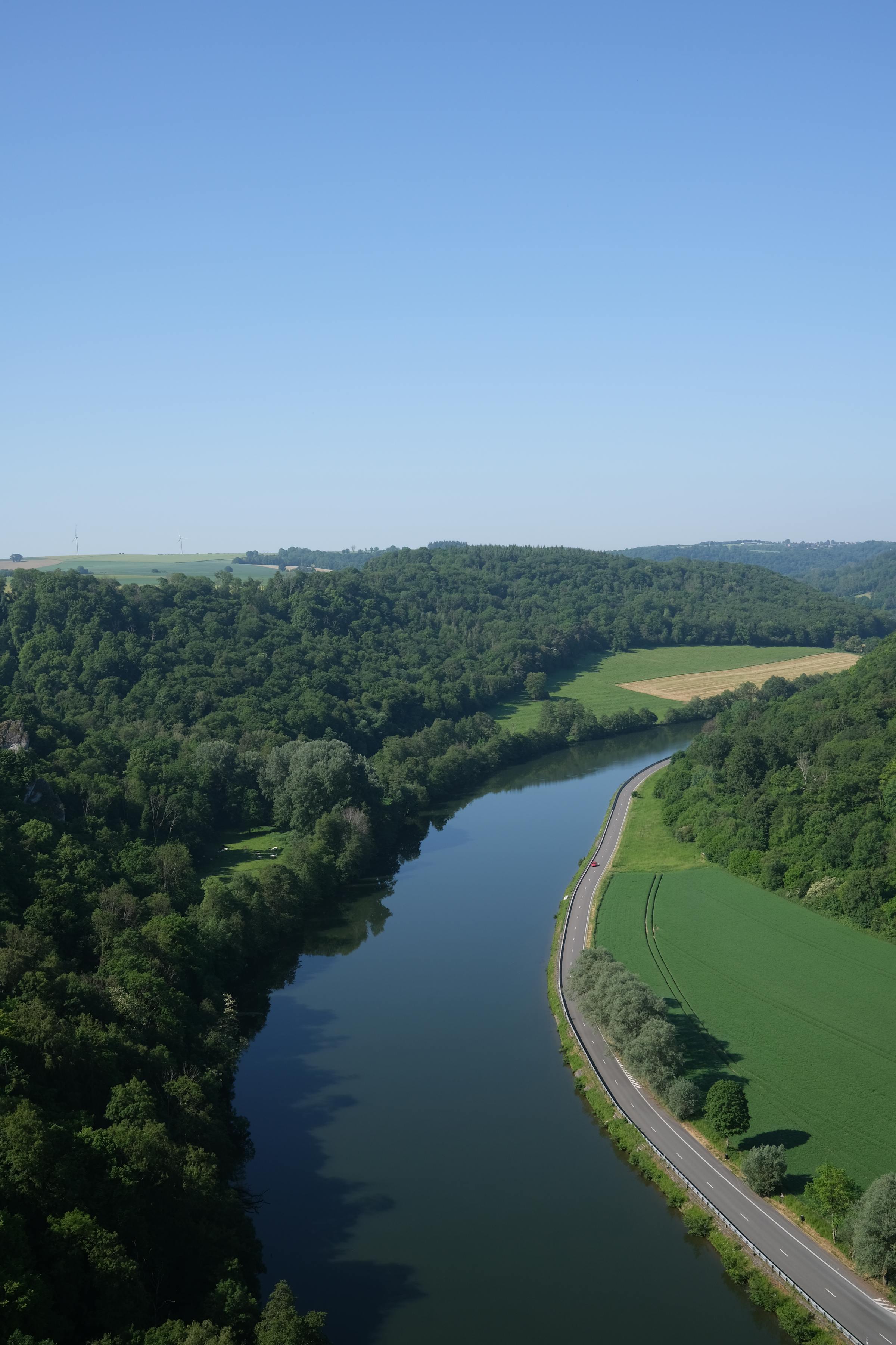 Aerial view of a river curving through wooded hills and fields in Wallonia, Belgium, with a road following its bank.