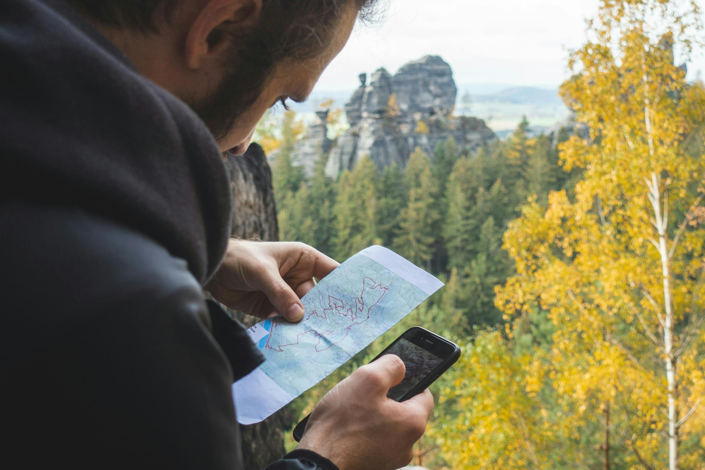 A hiker cross-checks a paper map against a phone in a forested, rocky landscape.