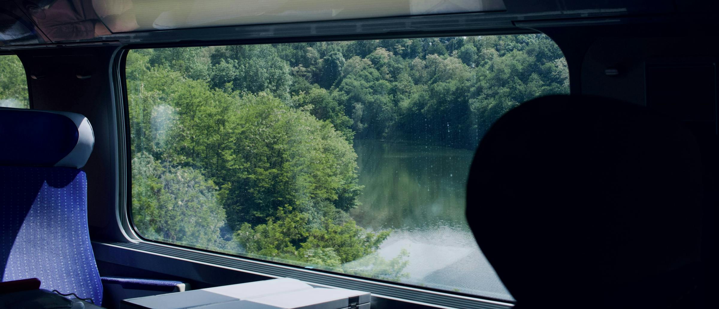 A wooded lake seen through a train window from a blue upholstered carriage seat.