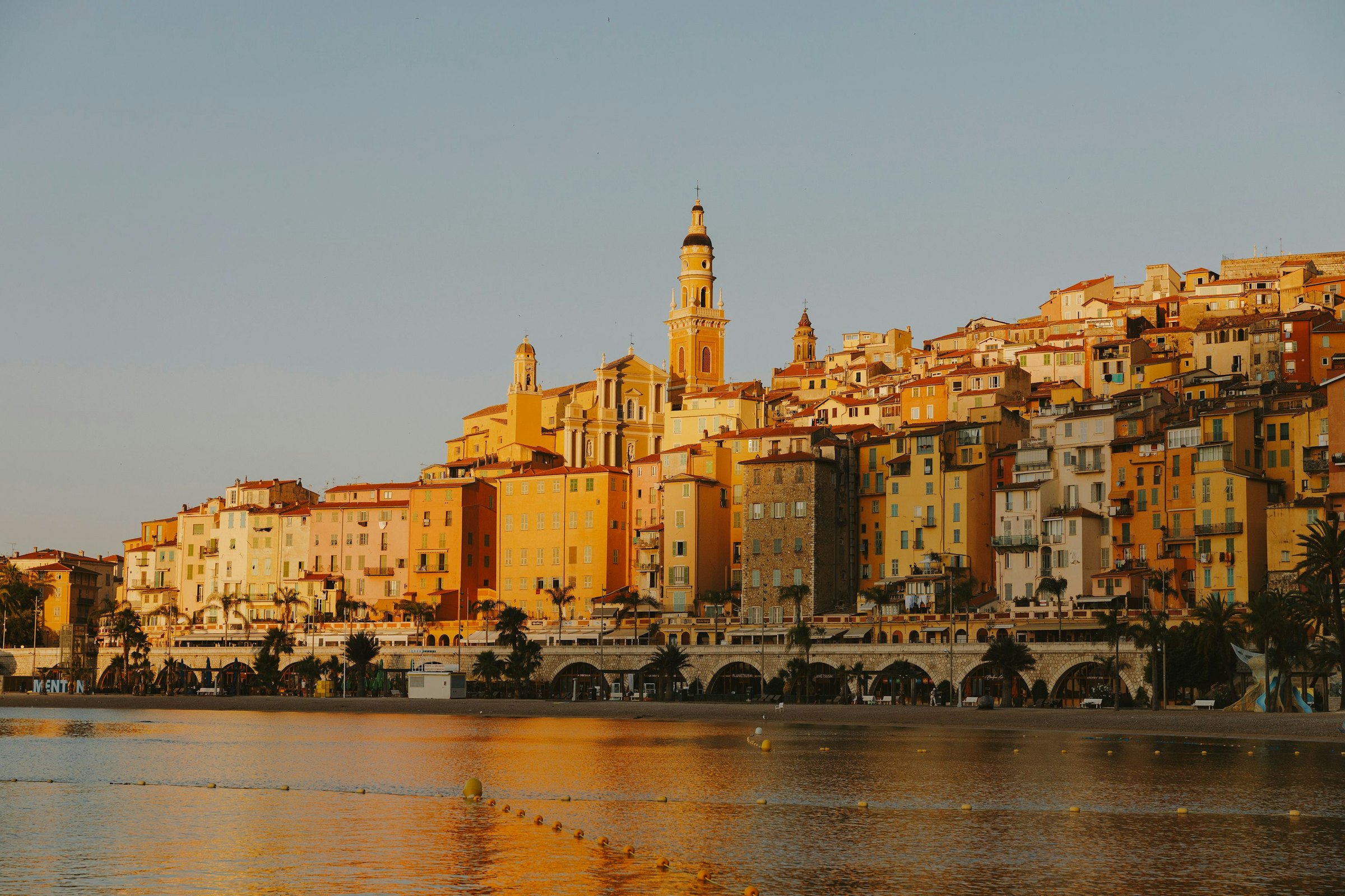 Warm sunlight illuminates the colorful, tiered hillside buildings and church tower of Menton, France, rising above the sea.