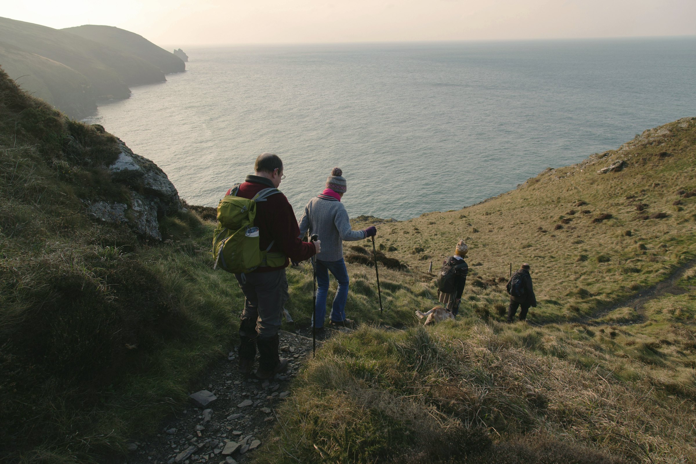 A group of hikers with trekking poles walks down a grassy coastal path overlooking cliffs and the sea.