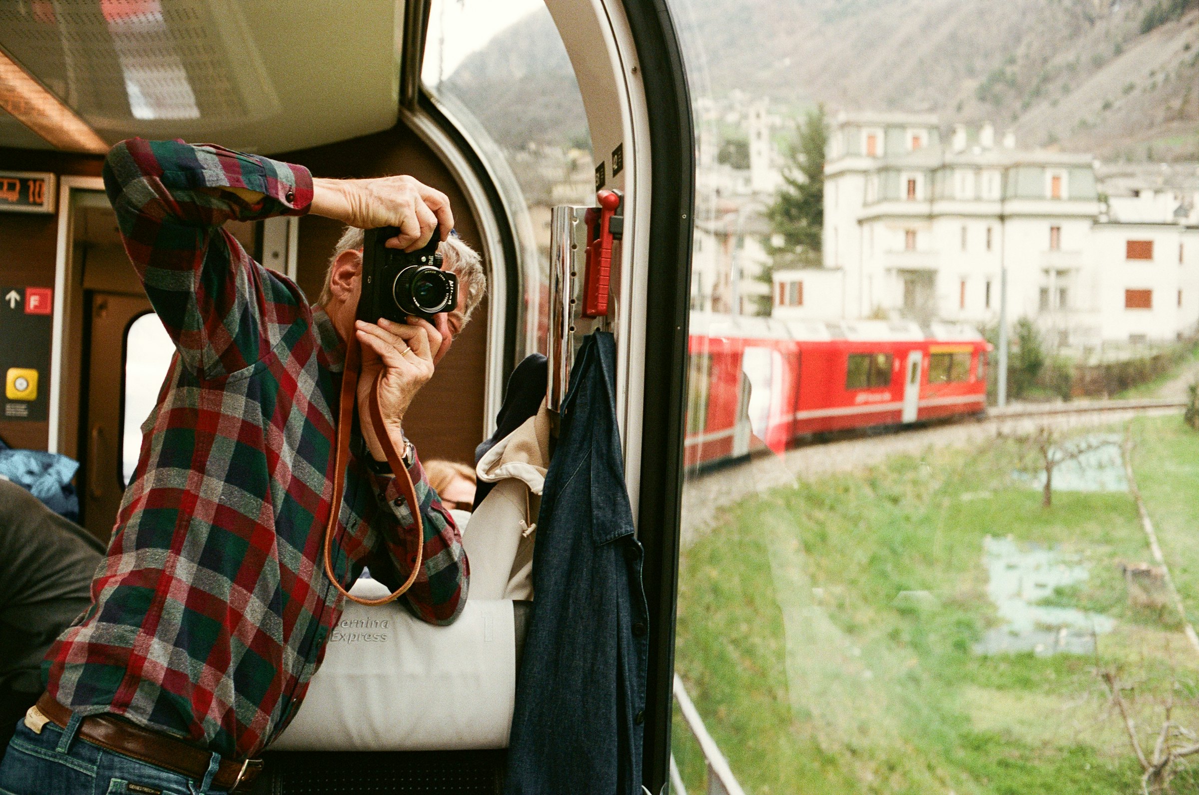 A man in a plaid shirt takes a camera photo inside a Bernina Express train, capturing the red carriage rounding a curve past Swiss village buildings.