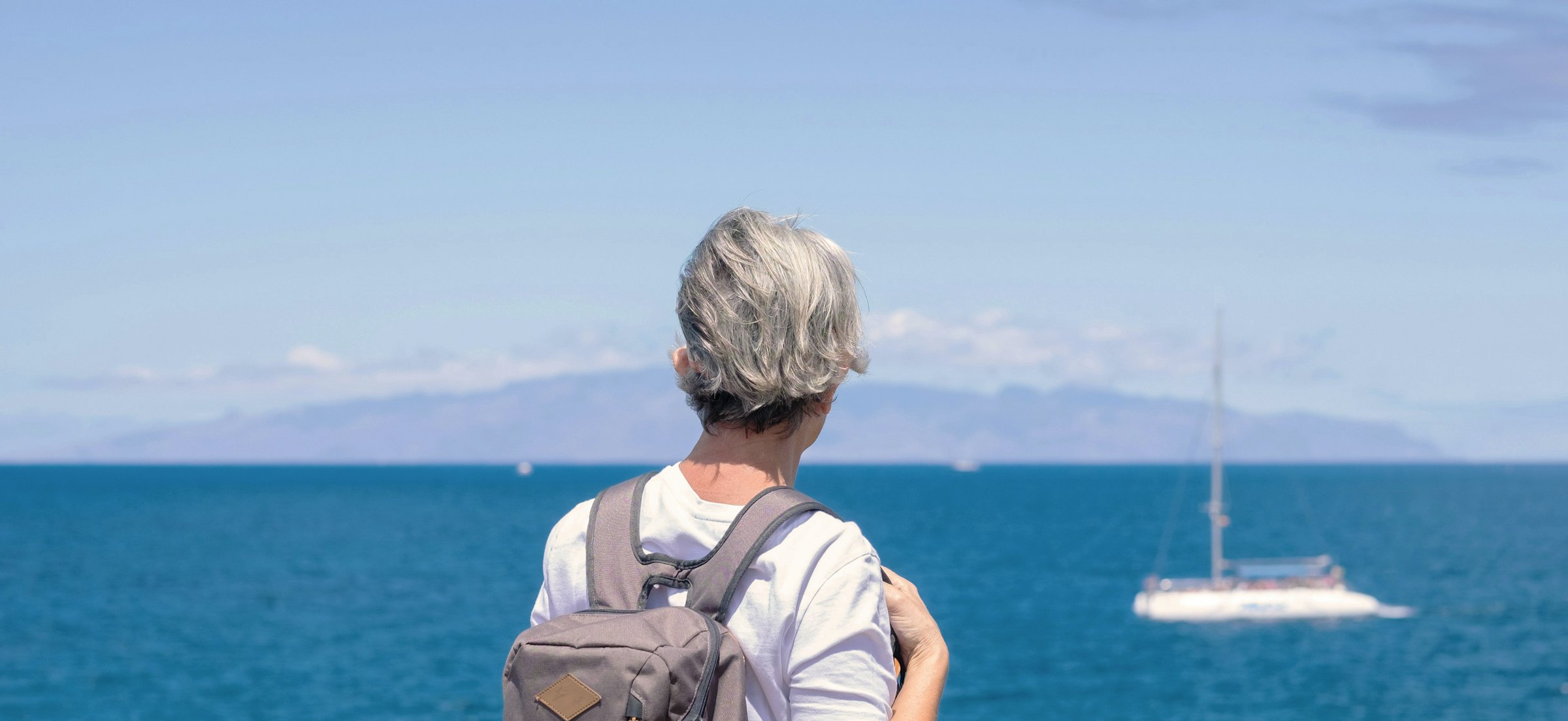 A woman with short grey hair and a backpack stands facing away, looking at a sailboat in the bright blue sea.