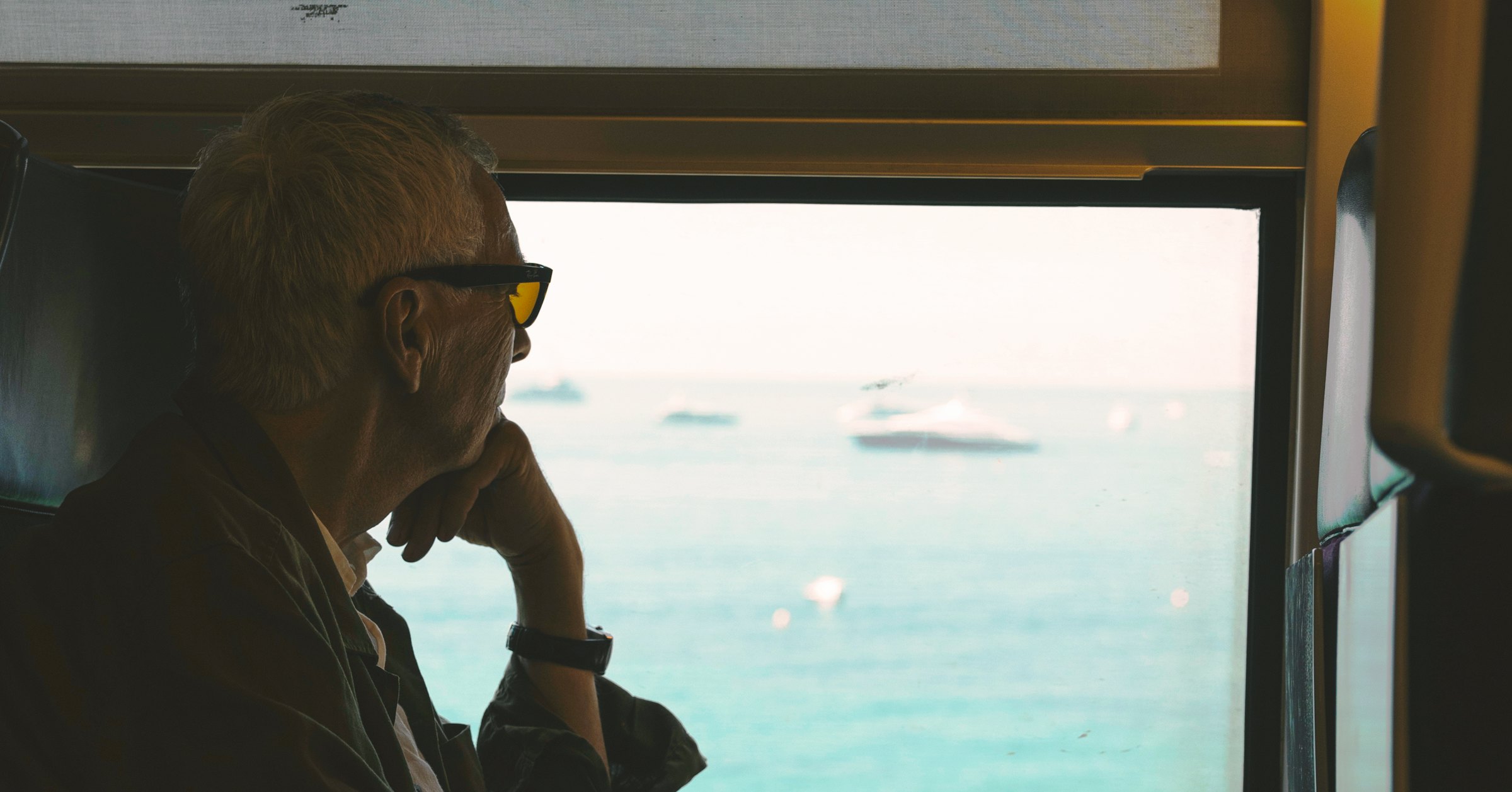 A silhouetted man in sunglasses looks out a train window at boats scattered across the French Riviera sea.