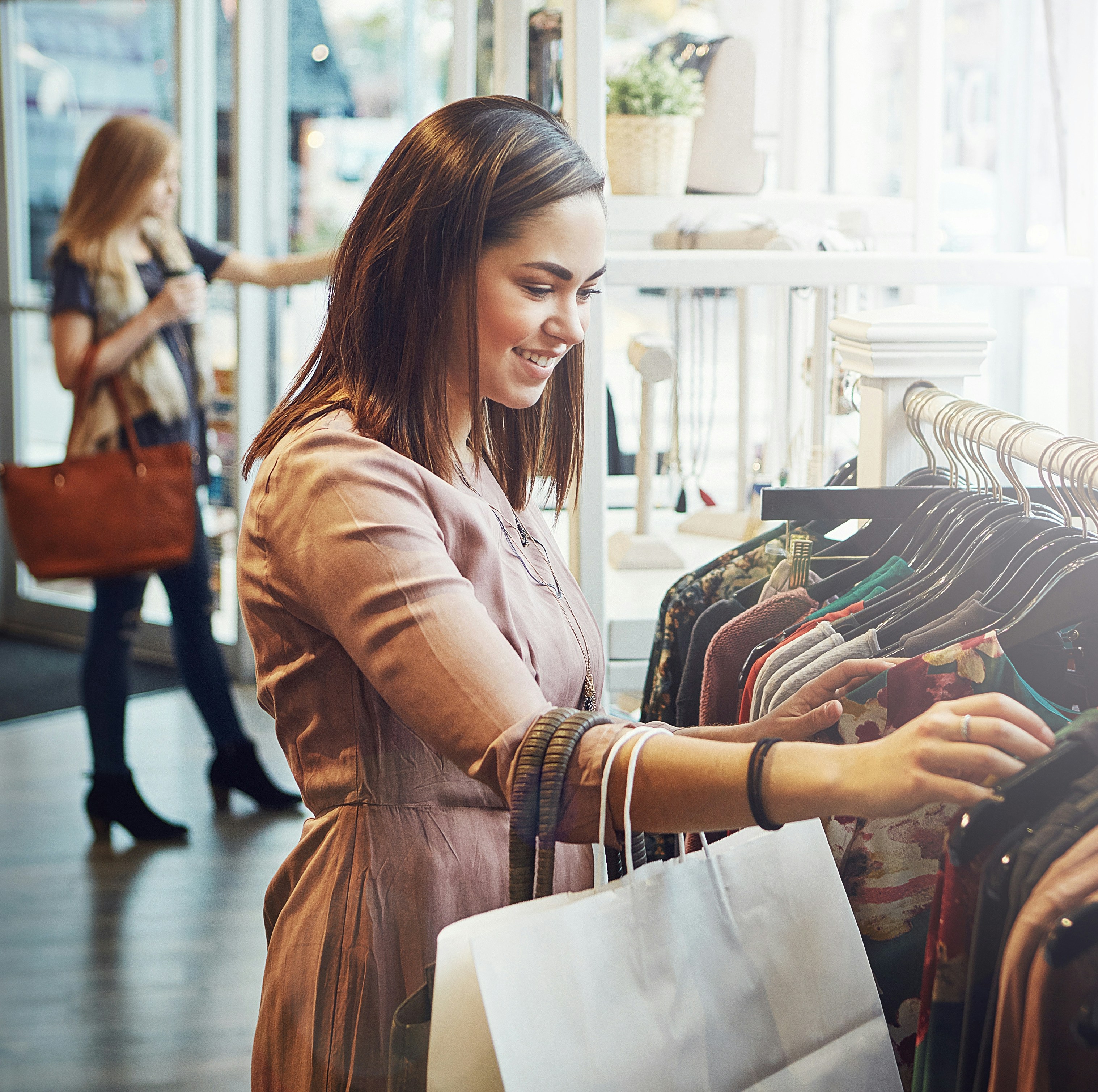 A smiling woman shopping for clothes in a bright, stylish store. She is browsing through a rack of clothing, holding shopping bags in her hand. Another shopper in the background is walking with a handbag and a drink, creating a bustling yet relaxed retail atmosphere.