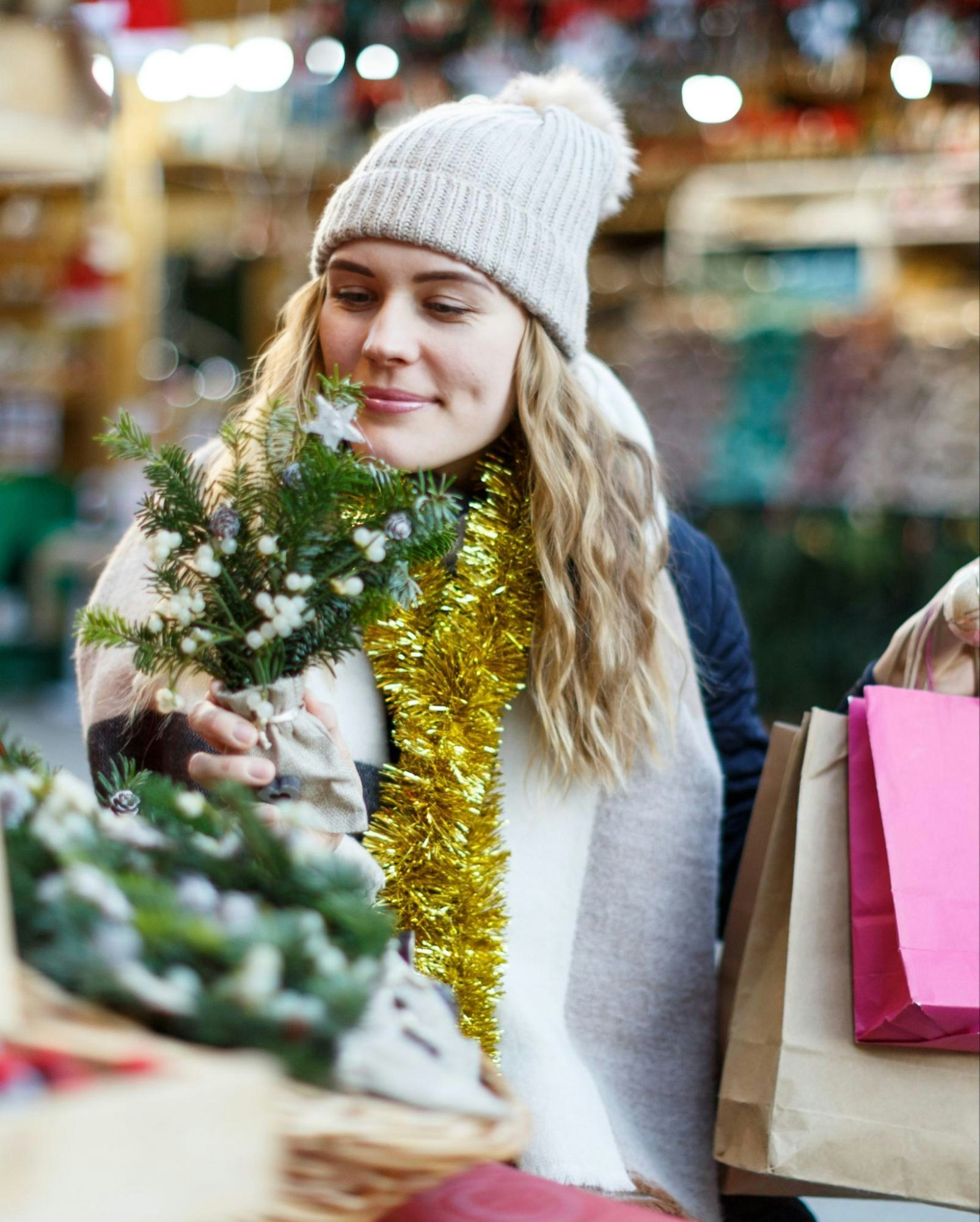 A woman wearing a knit hat and gold tinsel scarf shopping at a Christmas market, holding a small decorative tree with holiday decorations in the background.
