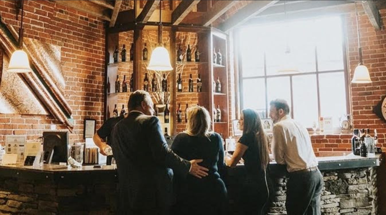 A group of people gathered at a rustic bar with exposed brick walls, wooden beams, and shelves stocked with wine bottles, creating a warm and inviting atmosphere.