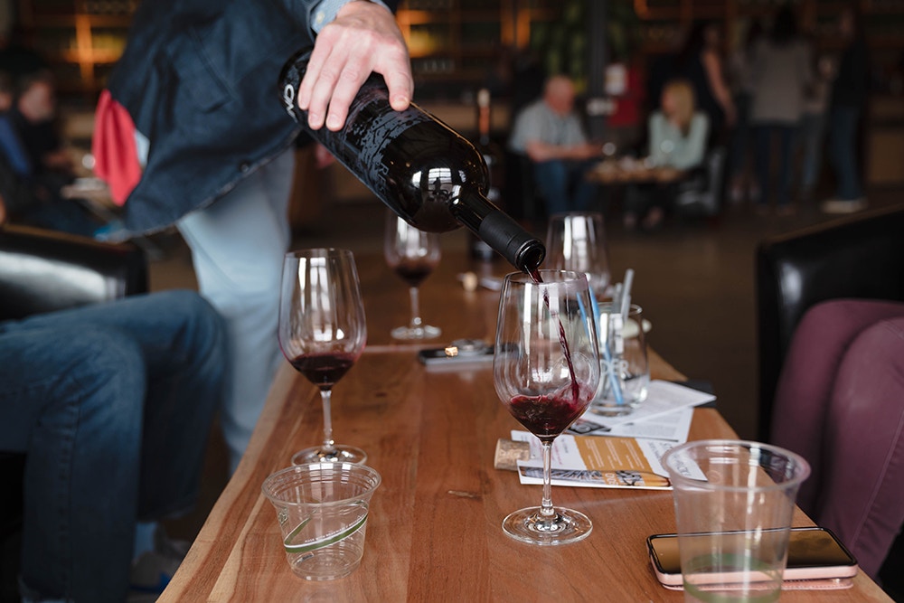 A close-up shot of a person pouring red wine into a glass at a social gathering in a dimly lit lounge. Several wine glasses, plastic cups, a smartphone, and menus are scattered on a wooden table, with a blurred background of people mingling.
