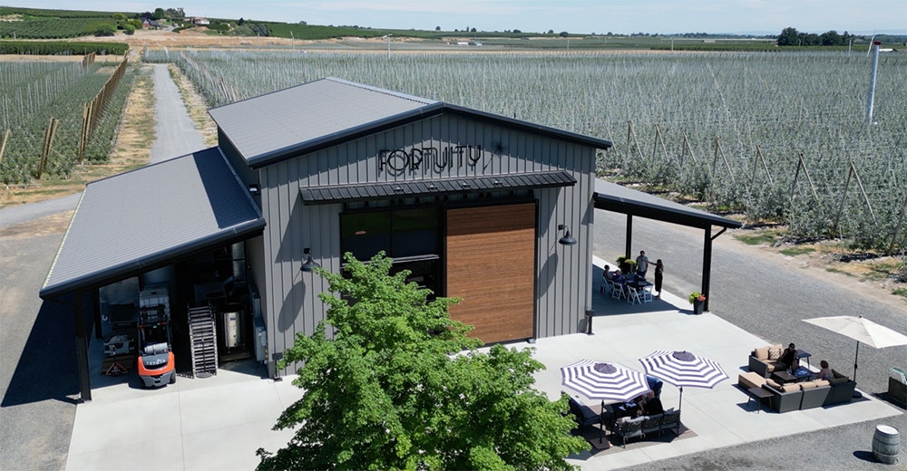 An aerial view of a modern winery building named 'Fortuity,' surrounded by lush vineyards. The structure has a sleek metal roof and outdoor seating areas with striped umbrellas, while visitors enjoy the space.