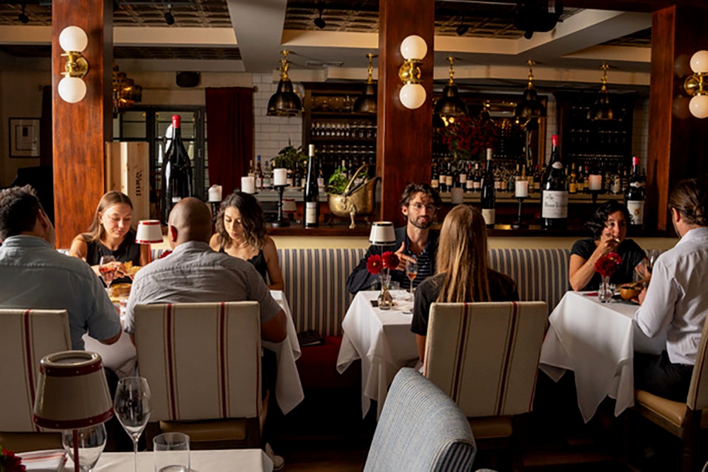A warmly lit, upscale restaurant with guests enjoying fine dining and wine. The setting features elegant décor, plush striped chairs, white tablecloths, and a well-stocked bar with large wine bottles in the background.